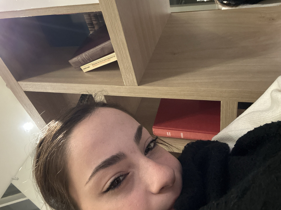 A person lies on their side, next to a wooden bookshelf. On the top shelf sit stacked books. The lower shelf contains a red-covered book. The person's face is close to the camera, and they appear to be resting. A black, fuzzy fabric is visible near their face. The wall behind them is white.