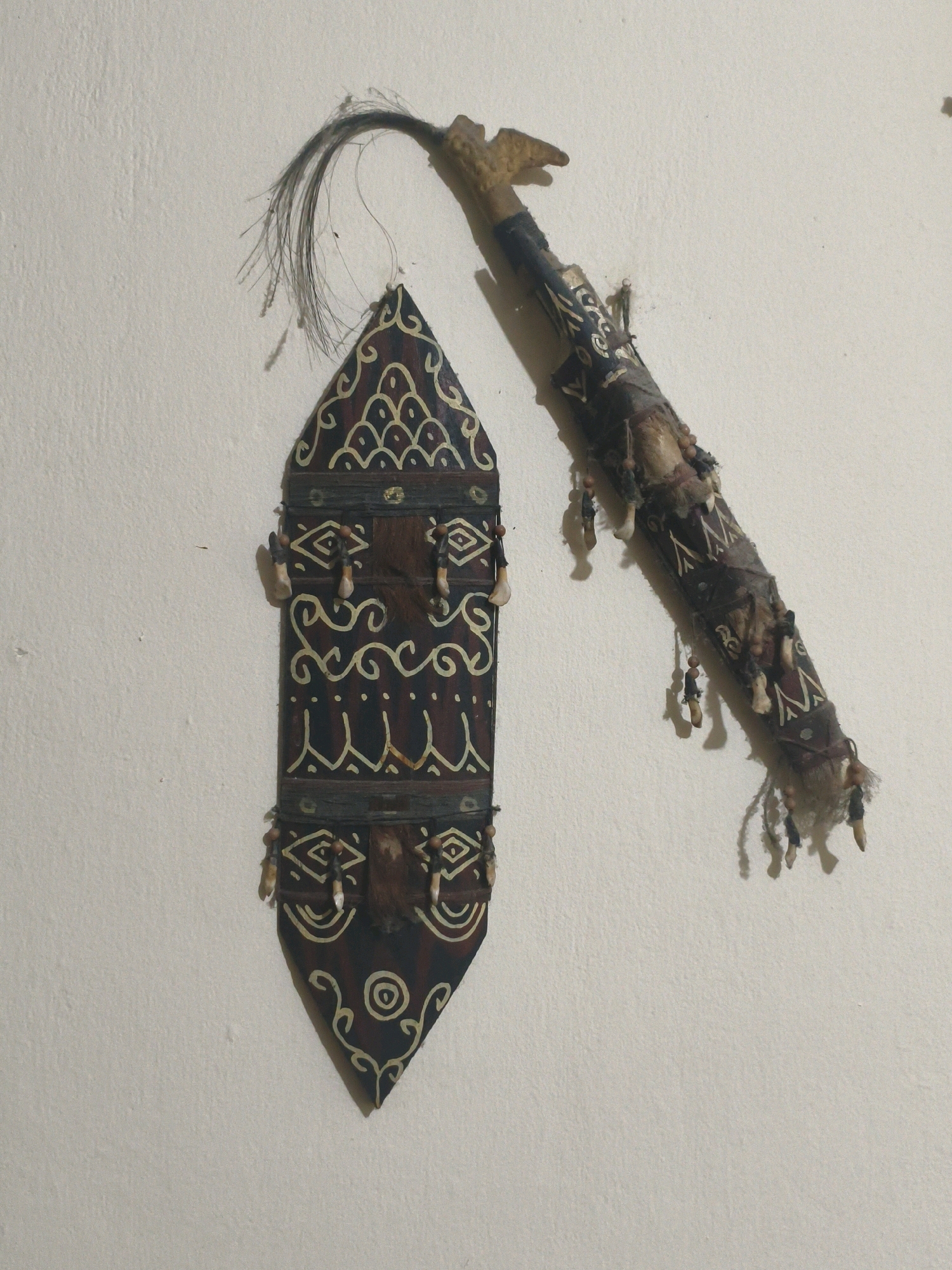 The image shows two tribal artifacts hanging on a textured, off-white wall. The artifact on the left is a shield-shaped object with intricate dark red and cream-colored geometric and curvilinear patterns. It has what appear to be small teeth or bone fragments and tufts of fur attached along its sides. The artifact on the right is a long, cylindrical object, possibly a scabbard or spear, also adorned with fabric, fur, teeth-like elements, and dangling beads. A tuft of dark hair extends from the top of this object. The lighting suggests an indoor setting. The artifacts are decorative and appear to be of cultural or ceremonial significance. This scene is not located in Bogor, Indonesia.