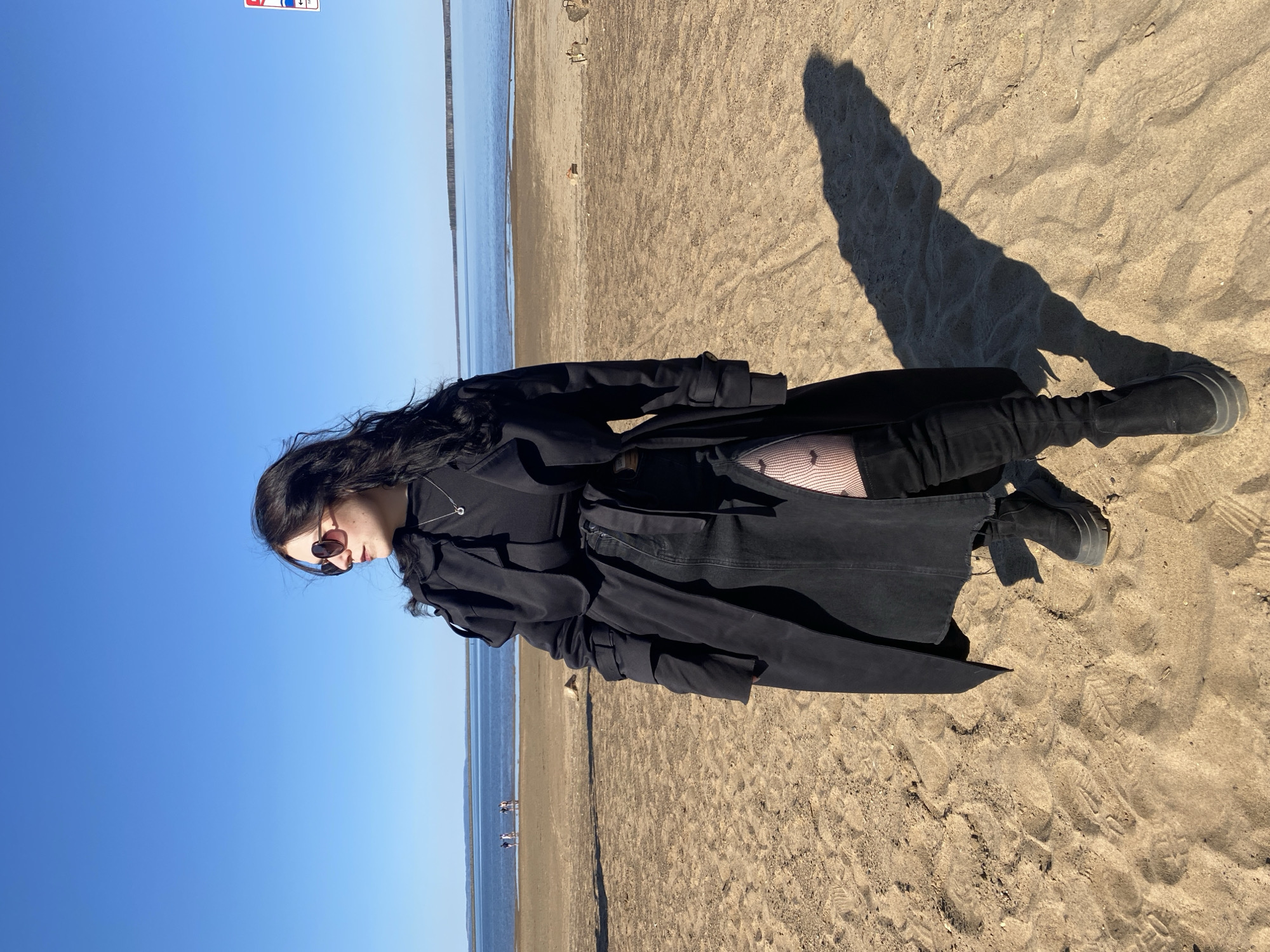 A woman in a black coat and boots stands on a sandy beach under a clear blue sky. She is facing left, and her shadow is cast on the sand to her right. The sea is visible in the background, and two figures can be seen walking along the beach in the distance. The location is the city of Chaikovsky, Russia.