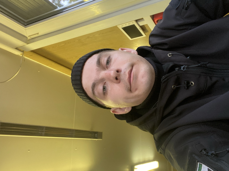 An adult male is centered in a close-up, chest-up indoor portrait. He has light skin, short hair visible beneath a black knitted beanie, and is wearing a black high-collared garment under a black tactical jacket. The jacket features multiple zippers and a patch on the left sleeve displaying vertical green, white, and dark grey/black stripes. He looks directly at the camera with a neutral expression. The indoor environment is illuminated by a warm, yellowish artificial light. The ceiling is light-toned, with two elongated fluorescent light fixtures visible, one prominent and another partially cropped on the left. The background consists of a light-colored wall on the left and a light wood-paneled wall on the right. A white-framed window or door is present on the right side of the frame, showing blurred outdoor elements beyond. Mounted on the wooden wall is a small, light-colored rectangular object with a dark screen, possibly a monitor or control panel, positioned above a partially visible red object. This scene is located in the city of Марфино, Russia.