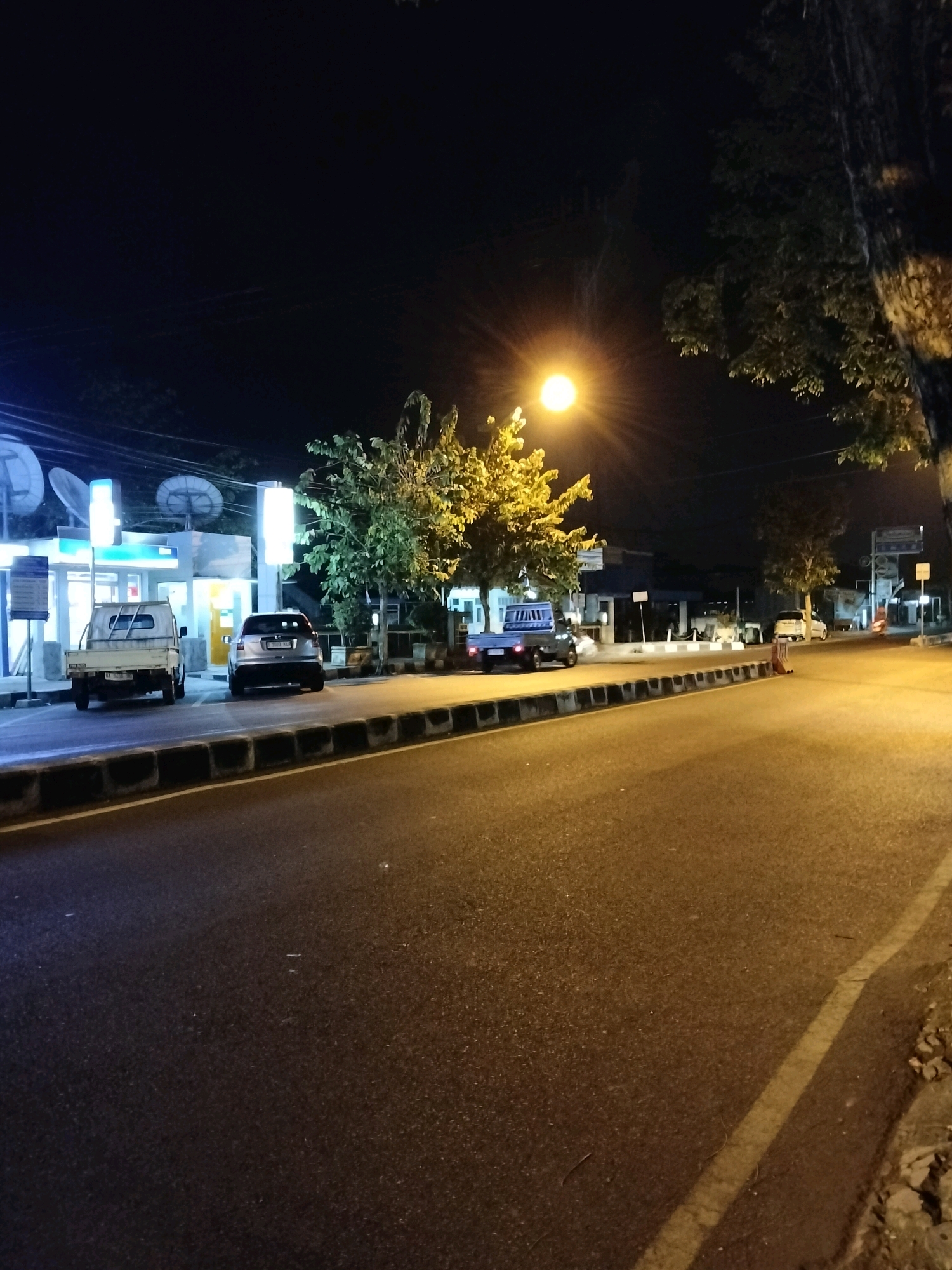 The image depicts a street scene at night in Purworejo, Indonesia. A street light illuminates a paved road with a sidewalk on the left. Several vehicles are parked along the sidewalk, including a white pickup truck and a silver SUV. In the background, a building with illuminated signage and satellite dishes is visible. Trees line the sidewalk, and the night sky is dark. The scene appears to be a commercial or residential area.