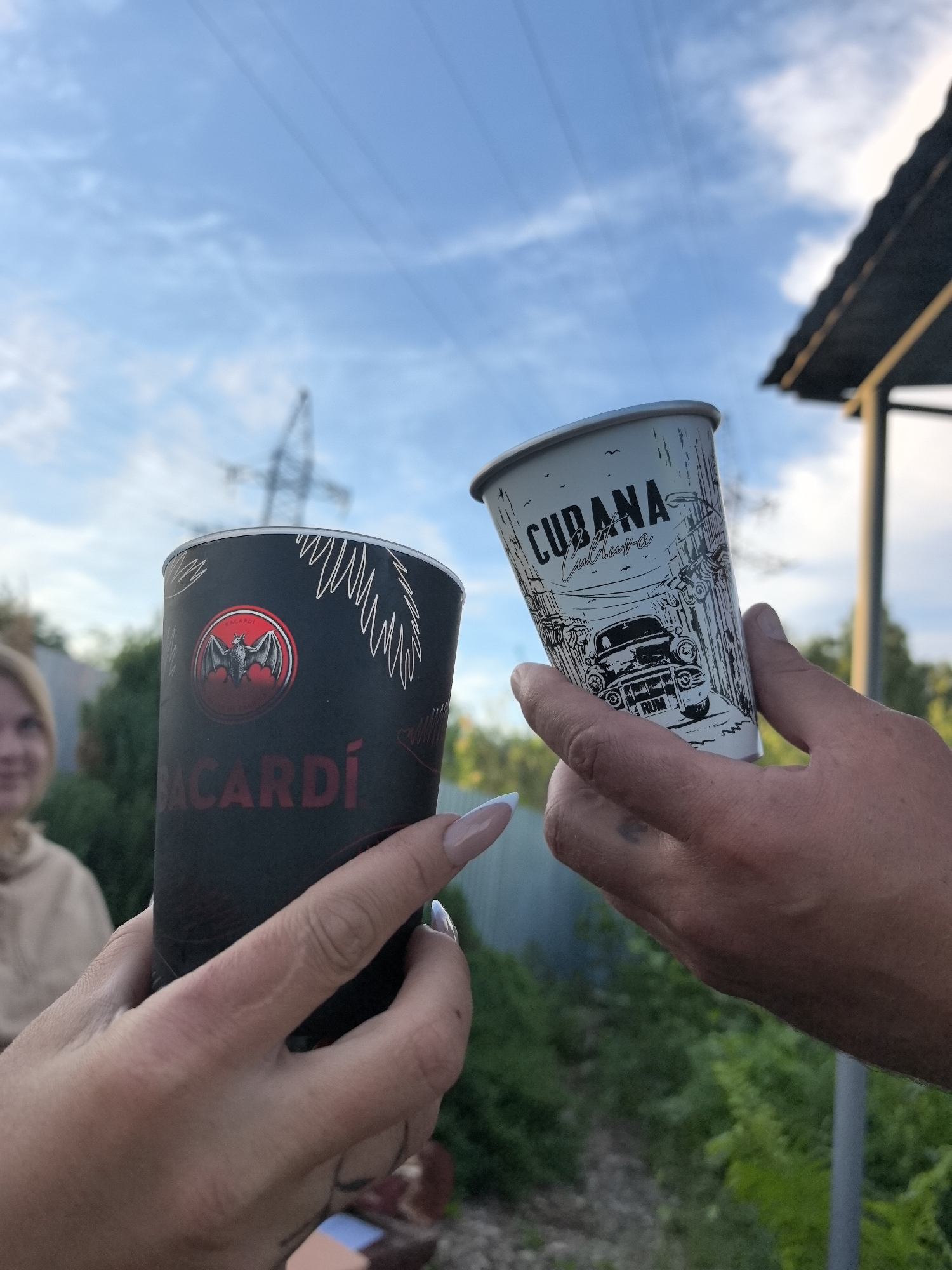 This outdoor, low-angle photograph, taken in Noviy, Russia, captures two individuals holding paper cups against a sky of scattered clouds and a backdrop of green foliage. The scene appears to be set in the late afternoon or early evening, characterized by soft, natural light.
In the foreground, a left hand with light-colored, manicured nails holds a black cup. This cup is adorned with a red circular Bacardí logo featuring a bat icon, and the word "BACARDÍ" in red text below it. White line art, resembling palm leaves, decorates the top part of the cup.
On the right, another hand holds a white cup. This cup features black line art depicting a vintage car within a street scene, possibly Havana-inspired. The text "CUBANA Cultura" is visible at the top of the design, with the word "RUM" placed near the car.
In the blurred background to the left, a blonde person, likely female, is partially visible, offering a slight smile. Behind them are green bushes and a corrugated metal fence. The mid-ground reveals prominent power lines stretching across the sky and a large, blurred power transmission tower. To the right, a dark-colored roof structure, possibly part of a shed or gazebo, is visible against more green foliage. The overall impression is of a casual, celebratory moment outdoors.