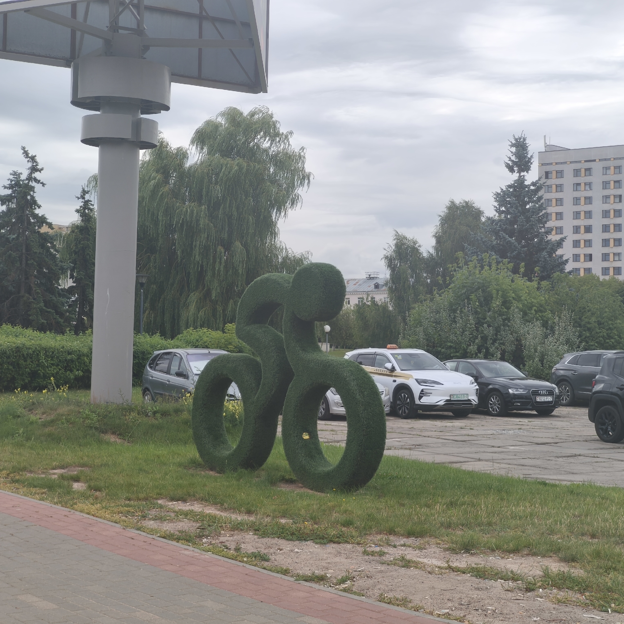 In Minsk, Belarus, on a cloudy day, a topiary sculpture resembling a cyclist in motion is positioned on a grassy area adjacent to a paved walkway. The sculpture, made of dense green shrubs, depicts a figure leaning forward, with circular elements suggesting wheels. Behind the sculpture, a parking lot is visible, containing several cars including a white taxi with a yellow roof sign, a dark sedan, and a dark SUV. In the background, a multi-story building with a grid of windows and trees, including weeping willows and evergreens, are present. A large, gray, cylindrical pillar with a metallic canopy is also a prominent feature in the foreground, located to the left of the topiary cyclist.