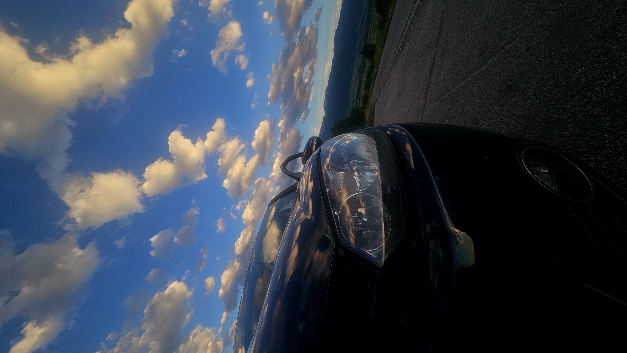 A low-angle, close-up shot of the front-left of a dark-colored car is positioned in the foreground, facing right. The car's headlight is clearly visible, reflecting the dramatic sky. The vehicle is parked on an asphalt road, which extends towards the right and into the distance. In the mid-ground and background, a vast landscape unfolds, featuring rolling hills or mountains covered in a layer of green vegetation. The sky above is a vibrant blue, dotted with numerous fluffy, white clouds that are illuminated by the setting sun, casting a warm glow. The overall scene suggests an outdoor setting, possibly a scenic overlook or a quiet road in a rural or mountainous area.