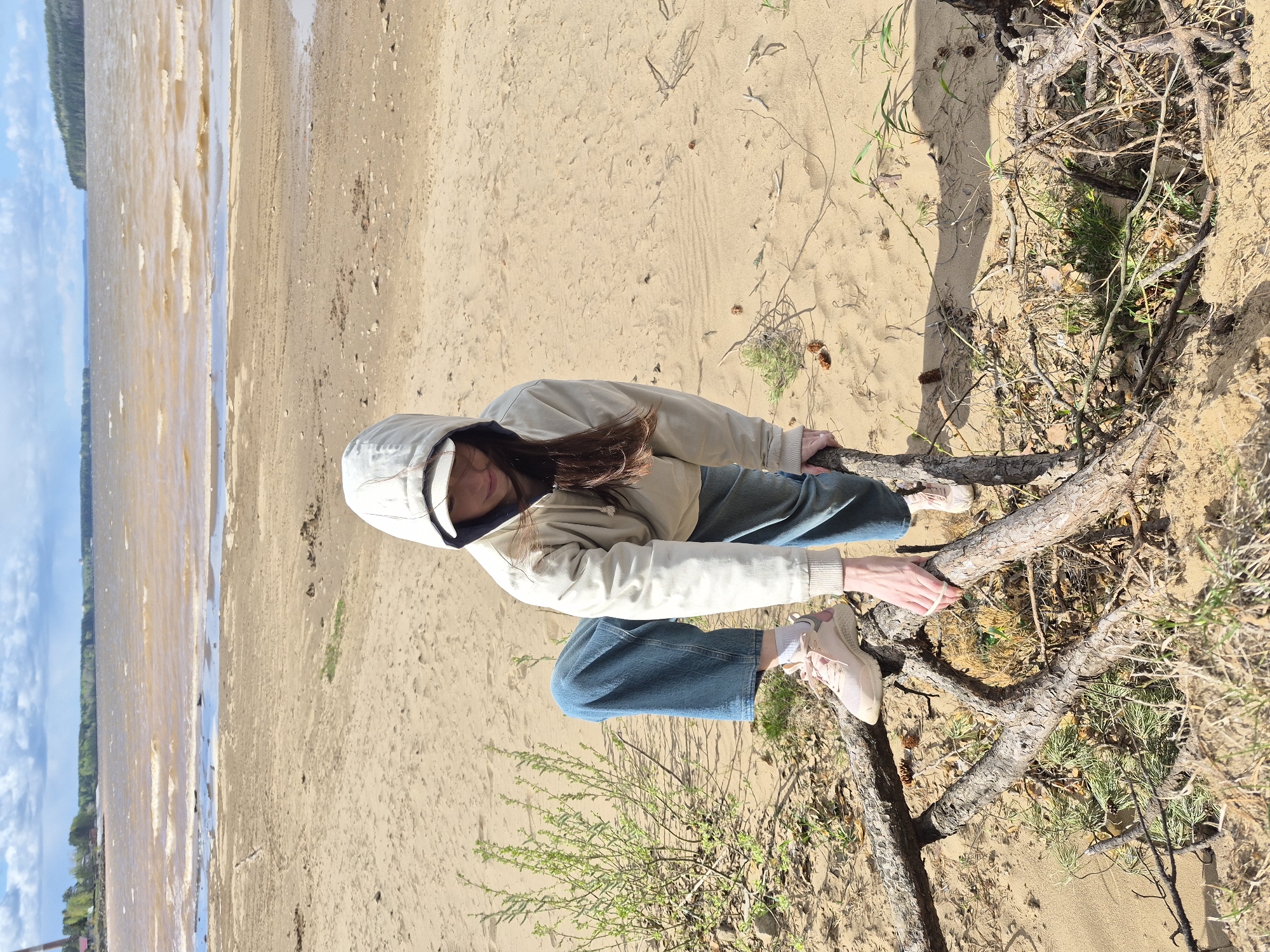 A woman in a hoodie and jeans sits amongst exposed tree roots on a sandy embankment at the edge of a body of water. The location is in the city of Chaikovsky, Russia, based on the photograph's origin. The water, possibly a river or lake, occupies the left side of the frame, with a strip of beach visible between the water and the embankment. The woman's feet and part of her legs are visible. She is holding onto a tree root with both hands. A small bush with green foliage is growing near the roots. The embankment has scattered dried leaves, twigs, and pinecones. The sky is visible above the water, showing a mix of clouds and blue sky.