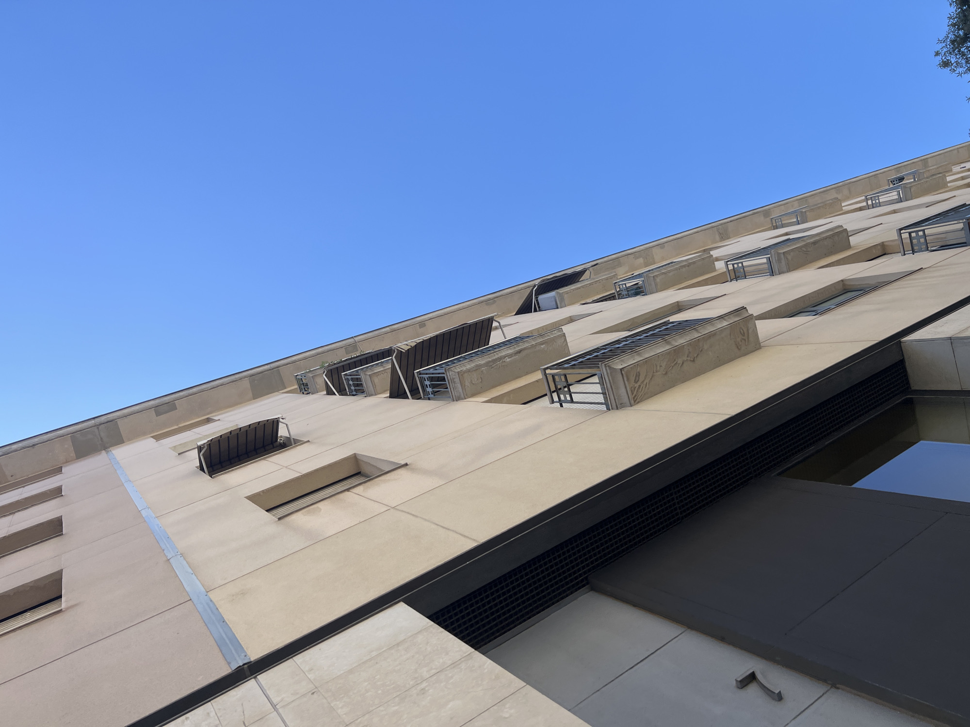 The image shows a low-angle, upward view of a cream-colored building with multiple balconies and windows against a bright blue sky. The building's facade is constructed with large, rectangular panels. Several balconies, each with a dark metal railing and a concrete base, are visible, staggered across the upper floors. Above some of the balconies, awnings are extended. A dark, large opening, possibly an entrance or a bay window, is visible in the lower left of the frame, with a textured metal grate above it. A small, angular metal fixture protrudes from the wall near this opening. The architecture suggests a modern urban setting. The clear blue sky indicates fair weather. While the location is stated to be Barcelona, Spain, no specific landmarks or contextual clues within the image definitively confirm this. There are no people or animals visible in the frame.