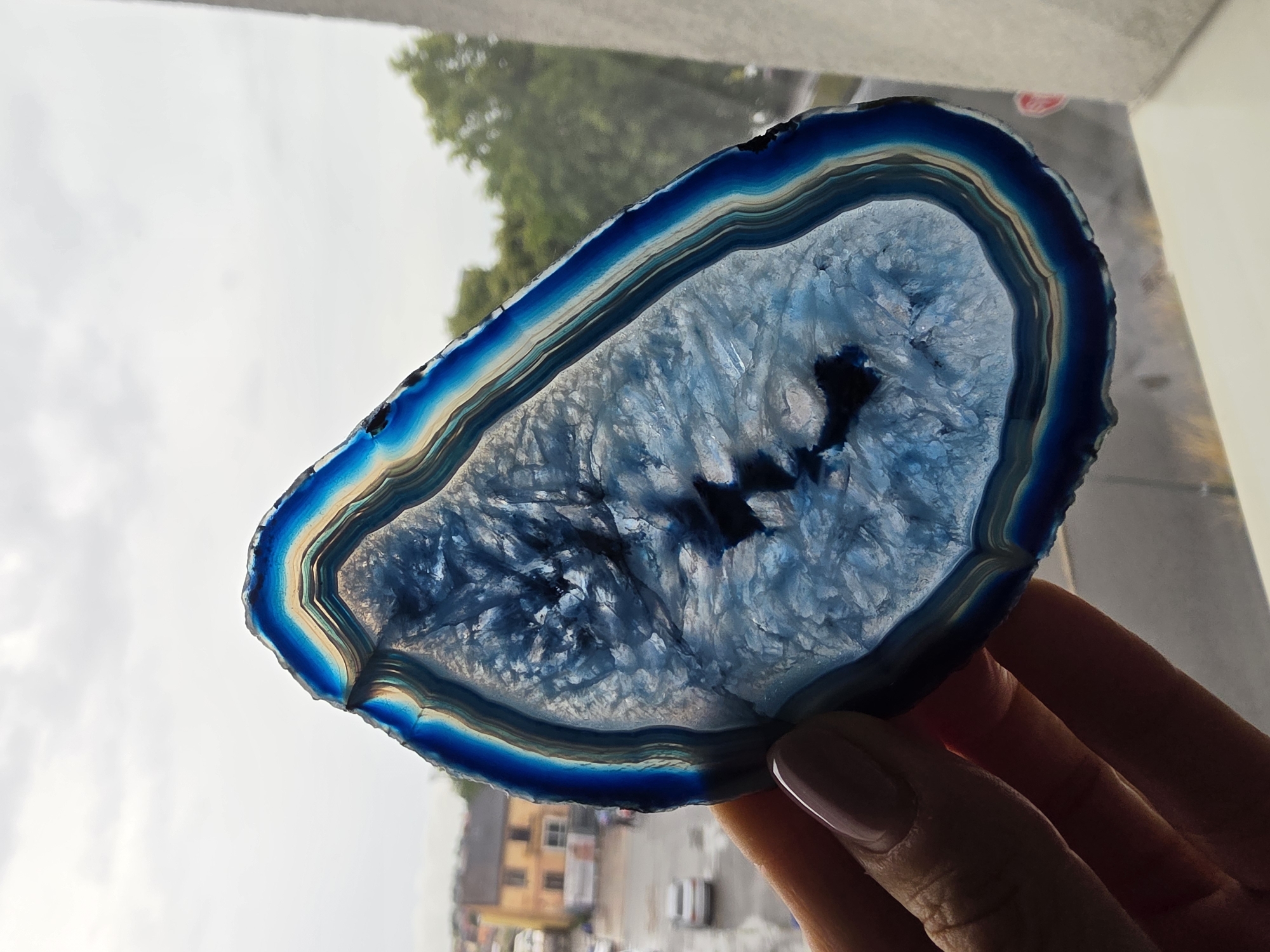 A sliced blue agate crystal is held in a person's hand in front of a window. The agate has a banded edge with layers of blue, white, and brown. Through the window, a street scene is visible, including buildings, cars, and trees. The sky is overcast. The image appears to have been taken outdoors. The specific location in the city of Poprad, Slovakia cannot be determined from the image.