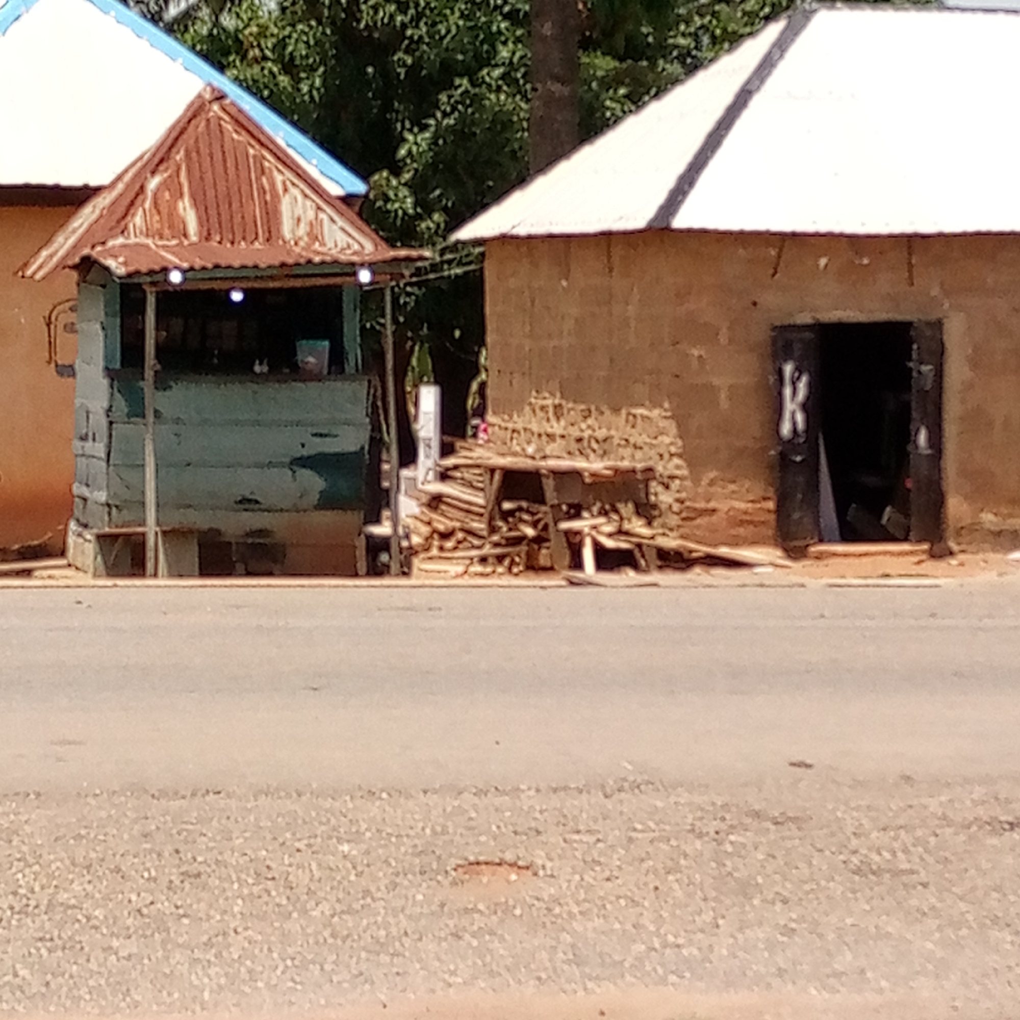 The image shows a street scene in Mararaba, Nigeria. In the foreground is a grey asphalt road. On the right side of the frame is a small, rectangular, mud-brick building. The building's open doorway reveals a dark interior. A white letter "K" is painted on the doorframe. Adjacent to the building is a haphazard pile of wooden planks and sticks.
To the left of the woodpile is a small, blue-green wooden stall with a rusty corrugated metal roof. The stall has visible goods inside. To the left of the stall is a partial view of another mud-brick building with a blue and white roof.
The background is a blur of green foliage. The scene appears to be a commercial or residential area.