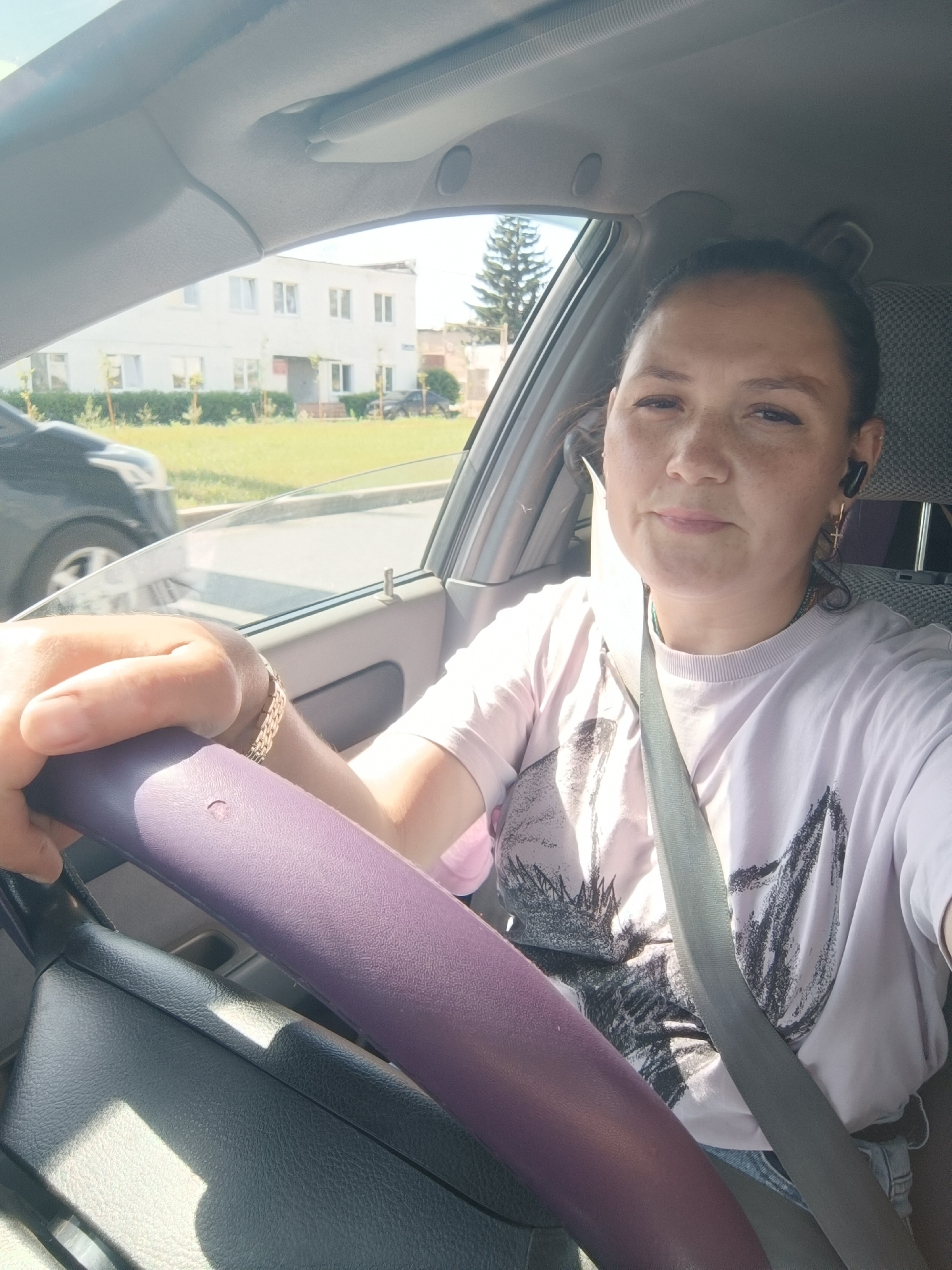The image shows a woman driving a car on a sunny day in Chaikovsky, Russia. The woman is wearing a light purple t-shirt with a graphic design and a seatbelt. She has an earpiece in her right ear and is wearing earrings. Her left hand is on the purple steering wheel. Outside the car, there is a building with windows and some trees. A dark-colored car is visible passing by in the background. The interior of the car, including the dashboard and windshield, is visible.