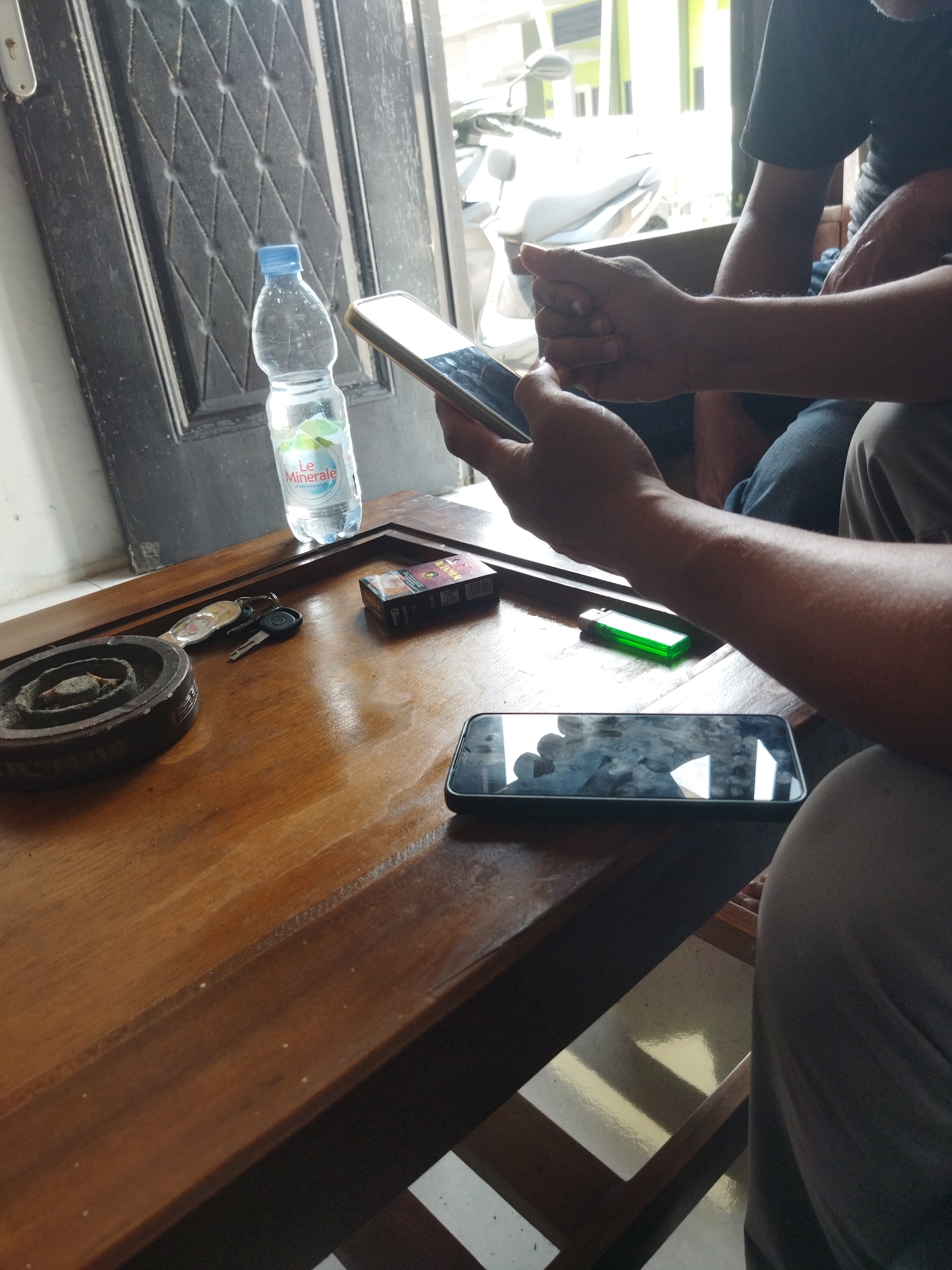 A medium shot captures a close-up view of a wooden table laden with various objects, with two individuals partially visible in the background. The table's polished surface reflects the ambient light, highlighting the rich grain of the wood. Positioned on the table are a bottle of Le Minerale bottled water, a pack of Dunhill cigarettes, a set of keys, a dark-colored ashtray with cigarette butts, a green lighter, and two smartphones. One smartphone, held by a person whose arms and hands are in focus, is angled towards the viewer, its screen displaying a mirrored reflection of the surroundings. Another smartphone rests face up on the table, its screen also reflecting the scene. The individuals in the background are indistinct, one appearing to be wearing a dark t-shirt and the other partially visible in gray trousers. The environment suggests an indoor setting with a door adorned with a diamond-patterned design to the left and a glimpse of what appears to be a motorcycle and bright outdoor light in the background. The overall impression is that of a casual gathering, possibly in a public space like a cafe or waiting area in Suka Sari, Indonesia, given the presence of the local brand of bottled water.