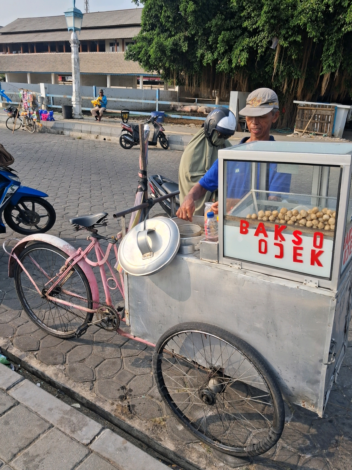 An outdoor scene in the city of Surakarta, Indonesia, shows a street vendor's cart as the central focus. The cart, constructed of metal, is a mobile food stall, with a glass display case at the front. Inside the case, numerous spherical meatballs are visible, and red lettering on the glass reads "BAKSO OJEK." A man wearing a baseball cap and a blue shirt stands behind the cart, seemingly attending to it. His left hand is inside the display case, and his right hand is near a plastic bottle.
Beside the cart, a pink bicycle with a black seat is parked. The bicycle is a three-wheeled model, with two wheels in the back and one in the front. A blue motorcycle is parked to the left of the bicycle, and another motorcycle is partially visible in the background to the right. The ground is paved with interlocking stone pavers. In the background, a building with a dark roof and light-colored walls is visible, and large trees with hanging roots provide shade. A street lamp with a blue top stands near the building. A man in a blue shirt is sitting on a bench in the background, holding a yellow object.