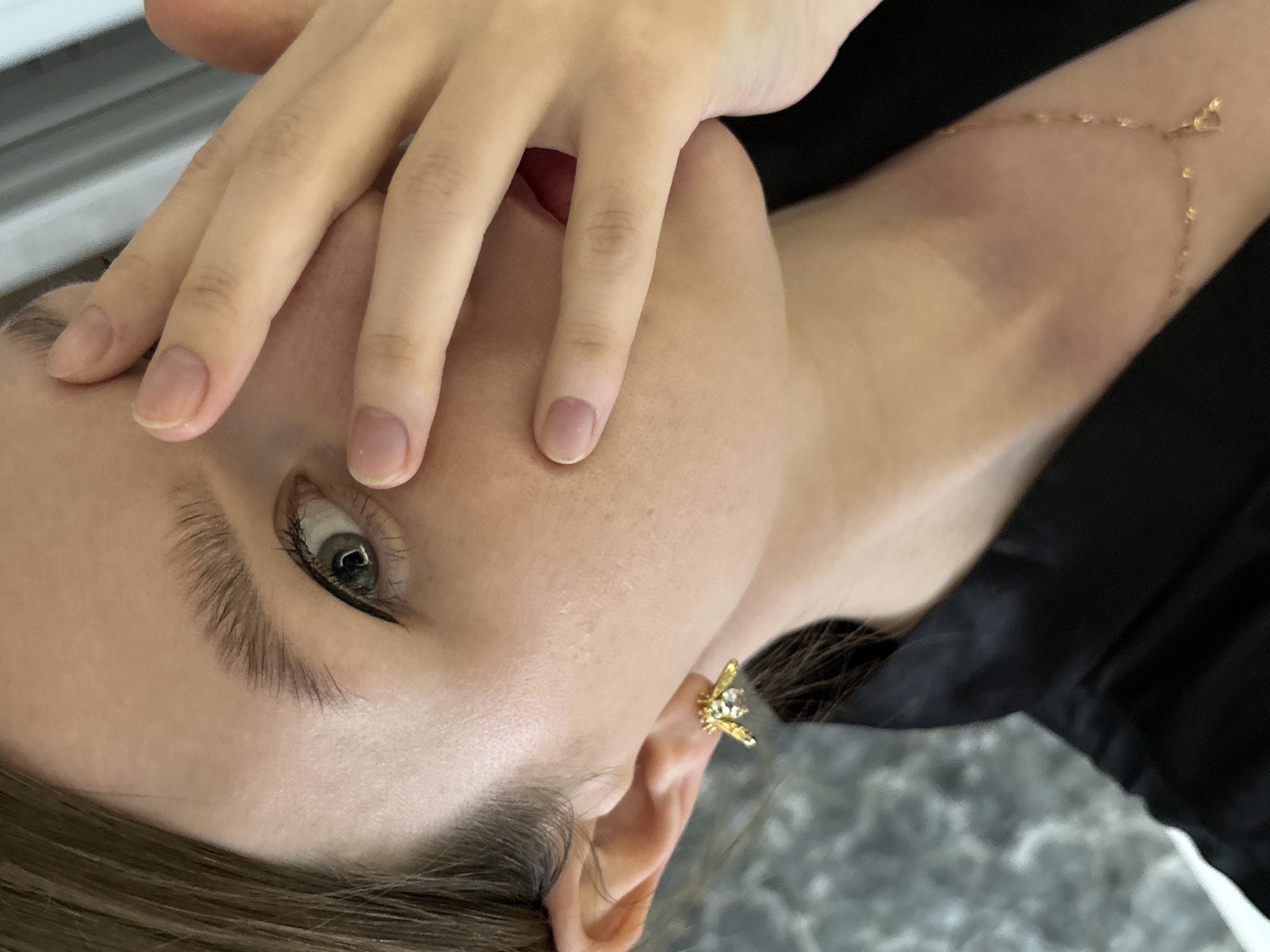A close-up, angled view of a light-skinned person covering part of their face with their hand. Their left eye is visible, enhanced with black eyeliner. They are wearing a gold-colored earring shaped like a bee. A delicate gold chain adorns their collarbone/upper arm area. The person is wearing a black garment. The background appears to be a light-colored surface with a textured pattern. It is not possible to ascertain the location in Izhevsk, Russia, from the provided image.