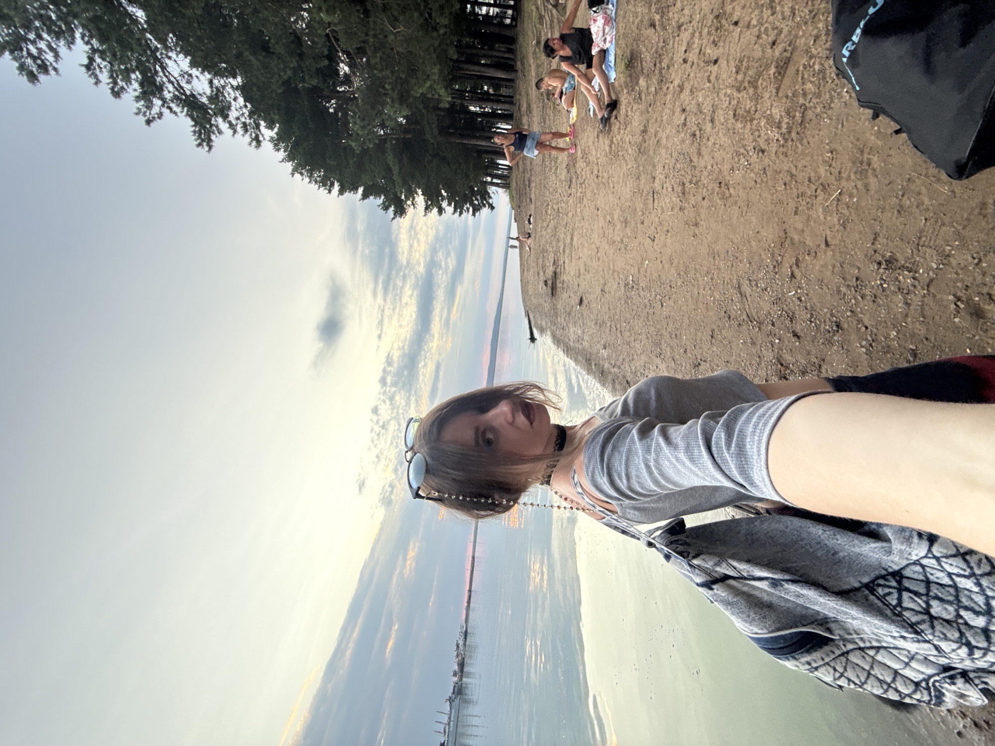 A medium-shot, eye-level, selfie-style video captures a young woman with dark hair and bold makeup standing on a sandy beach. She wears a gray, long-sleeved, off-the-shoulder shirt and dark shorts, and a beaded chain is attached to her sunglasses, which are perched on her head. She carries a large patterned bag over her shoulder. The background shows a calm body of water with a distant industrial area visible on the horizon, and a dense line of pine trees bordering the beach to the right. Several other people are scattered on the beach, some lying down and others standing. The lighting suggests it is late afternoon or early evening, with a soft, diffused light illuminating the scene.