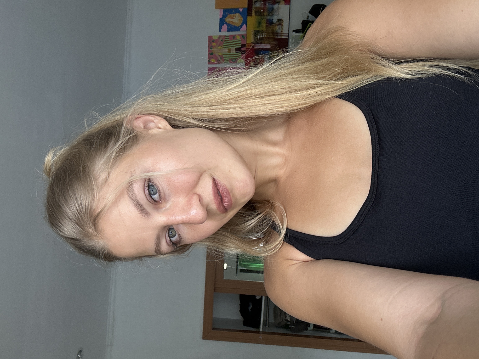 A close-up, chest-up shot features a young woman indoors, likely in a room in Kuta Selatan, Indonesia. She is the sole subject, looking directly at the camera with a calm, neutral expression or a slight, soft smile. She has long, blonde hair, which flows down her back and over her shoulders, and light blue eyes. She is wearing a black tank top.
The background consists of a plain light-colored wall, possibly white or light grey. On the upper right side of the frame, there are several colorful papers or drawings tacked to the wall, suggesting a personalized space. Below these, part of a wooden frame, possibly from a mirror or a window, is visible, reflecting a darker, indistinct area within the room. The lighting appears to be natural and diffused, indicating daytime. No text is visible in the image.