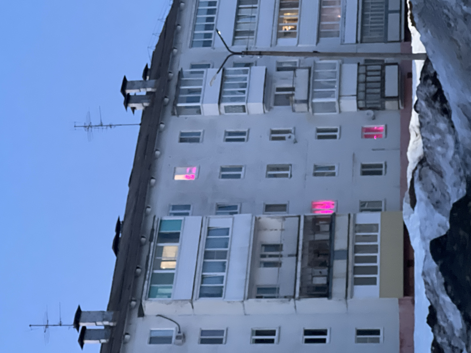This forensic description details a photographic capture of a multi-story residential building in the city of Noviy, Russia. The scene is illuminated by ambient twilight, indicated by a clear, pale blue sky, and features significant snow accumulation on the ground in the foreground.
The primary subject is a light-colored, multi-story apartment building, likely constructed from concrete or stucco, featuring a dark, gently sloping roof. Two prominent chimneys are visible on the left side of the roof, with a multi-element antenna structure positioned further right. The building facade is punctuated by numerous rectangular windows and several enclosed balconies with glazed panels.
Notably, three distinct windows emit a vibrant magenta-pink light from their interiors: one on an upper floor, one on a middle floor, and one on a lower floor. Other illuminated windows display standard warm-toned lighting. An external air conditioning unit is affixed to the building's left side, below an enclosed balcony.
A street lamp post, equipped with an illuminated fixture, stands vertically on the right side of the frame, partially obscuring the building. The foreground comprises irregular mounds of snow, interspersed with dark, bare ground or shadows, obscuring the lowest section of the building's foundation. No individuals, actions, or direct interactions are visible within the frame.