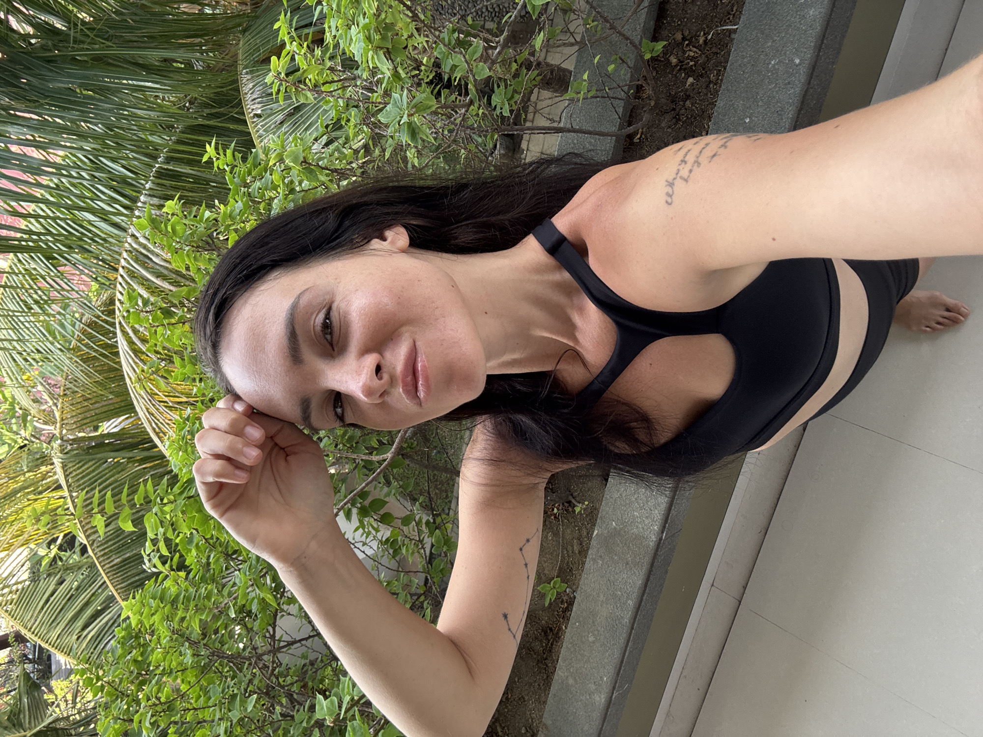 A woman with dark hair and fair skin is taking a selfie outdoors, with lush green foliage and palm fronds in the background. She is wearing a black sports bra and appears to be on a balcony or patio, with a tiled floor visible. There are tattoos on her left arm and shoulder. The lighting suggests it is daytime. The location is North Kuta, Indonesia.