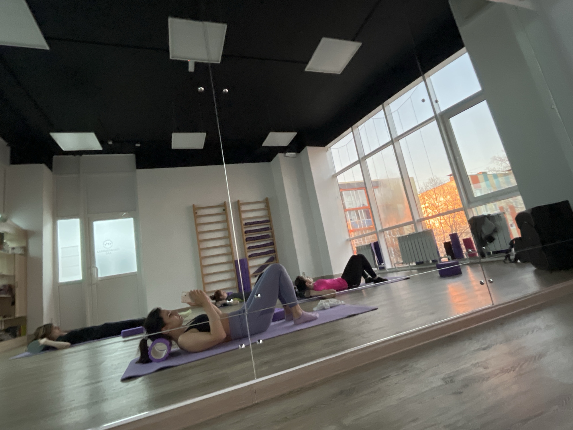 The image shows a mirrored view of a fitness studio in Petropavlovsk-Kamchatsky, Russia. Several women are lying on yoga mats, some using foam rollers. Exercise equipment, including wall bars, is present. Large windows reveal an outdoor view with buildings and trees. The room has a dark ceiling with rectangular lighting fixtures.