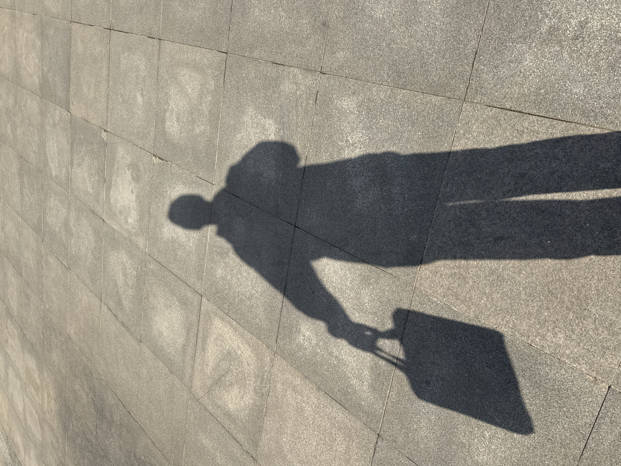 A shadow of a person pulling a wheeled suitcase is cast on a paved walkway or plaza. The paving consists of large, light gray, rectangular stones. The scene is located in Zhongtun, China.
