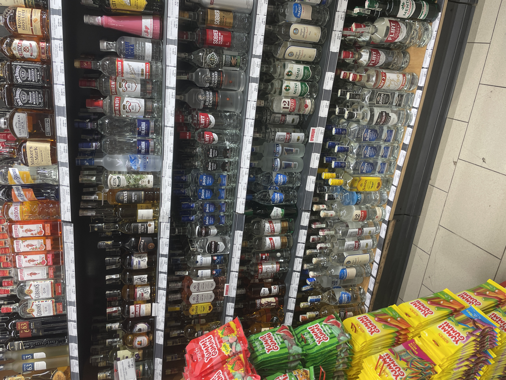 This image captures a well-stocked retail display of various alcoholic beverages and some confectionery, likely in a supermarket or liquor store in Ihtiman, Bulgaria. The primary subjects are numerous bottles of spirits arranged neatly on multi-tiered shelves.
The top shelves feature whiskies and brandies such as Monkey Shoulder, Chivas, Jack Daniel's, Gentleman Jack, and Maker's Mark. Below these are shelves predominantly displaying vodka brands including Stolichnaya, Smirnoff, Russian Standard, Flirt, Sobieski, Solut, Senses, Savoy, Alasca, Peristera, and Medusa. Gin brands like Gordon's, Beefeater, Gibsons, and Blue Brizard are also visible, alongside ouzo (Ouzo Plomari, Ouzo 12, Mini), and other spirits such as Menta (mint liqueur) and Mastika. Many bottles are smaller, indicative of miniature or single-serving sizes, especially among the ouzo.
At the bottom of the display, several bags of colorful candies are stacked, prominently featuring the brand "Roshen Yummi Gummi" in various flavors like Sour, Watermelon, and Rainbow Belts.
Price tags are visible throughout the shelves, displaying prices in Bulgarian Lev (лв), confirming the location context. A sign with "ПРОМОЦИЯ" (PROMOTION) is also present. The lighting is typical of an indoor commercial setting, with fluorescent lights illuminating the products. The flooring is tiled. No people are visible in the scene. The image provides a detailed view of the product selection and merchandising within this retail environment.