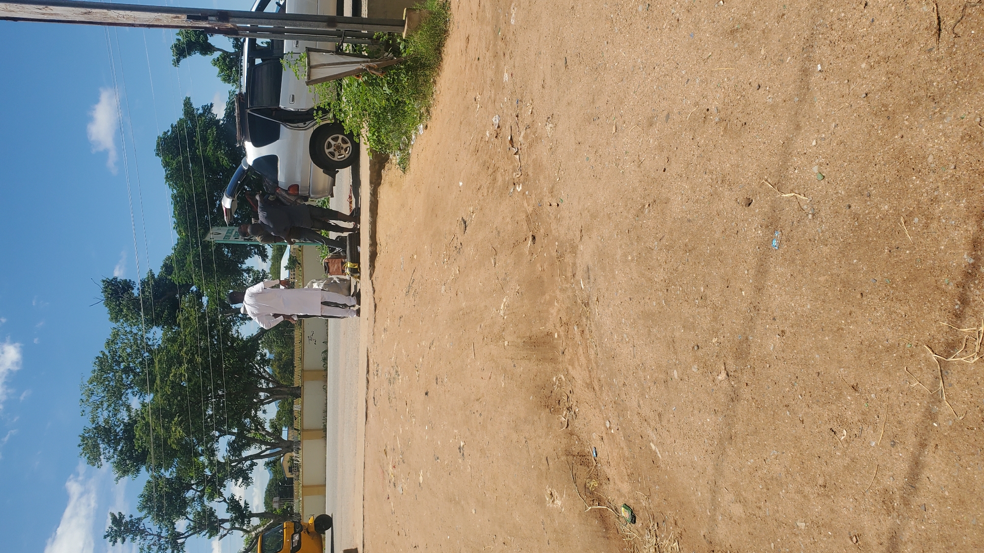 In Jalingo, Nigeria, near the edge of the road, a silver van with its rear hatch open is parked. Three men are gathered around the open hatch, seemingly engaged in an activity related to the van's cargo or contents. One man, standing slightly apart, is dressed in a white robe and pants. Another man, in dark clothing, leans into the open hatch. A third man, also in dark attire, stands between the two. A spare tire is visible on the ground next to the van. To the left, a yellow motorized tricycle is parked. The ground is dirt and sand, with scattered debris. In the background, lush green trees form a backdrop against a bright blue sky with scattered clouds. The scene appears to be in an outdoor, possibly urban or semi-urban, environment.