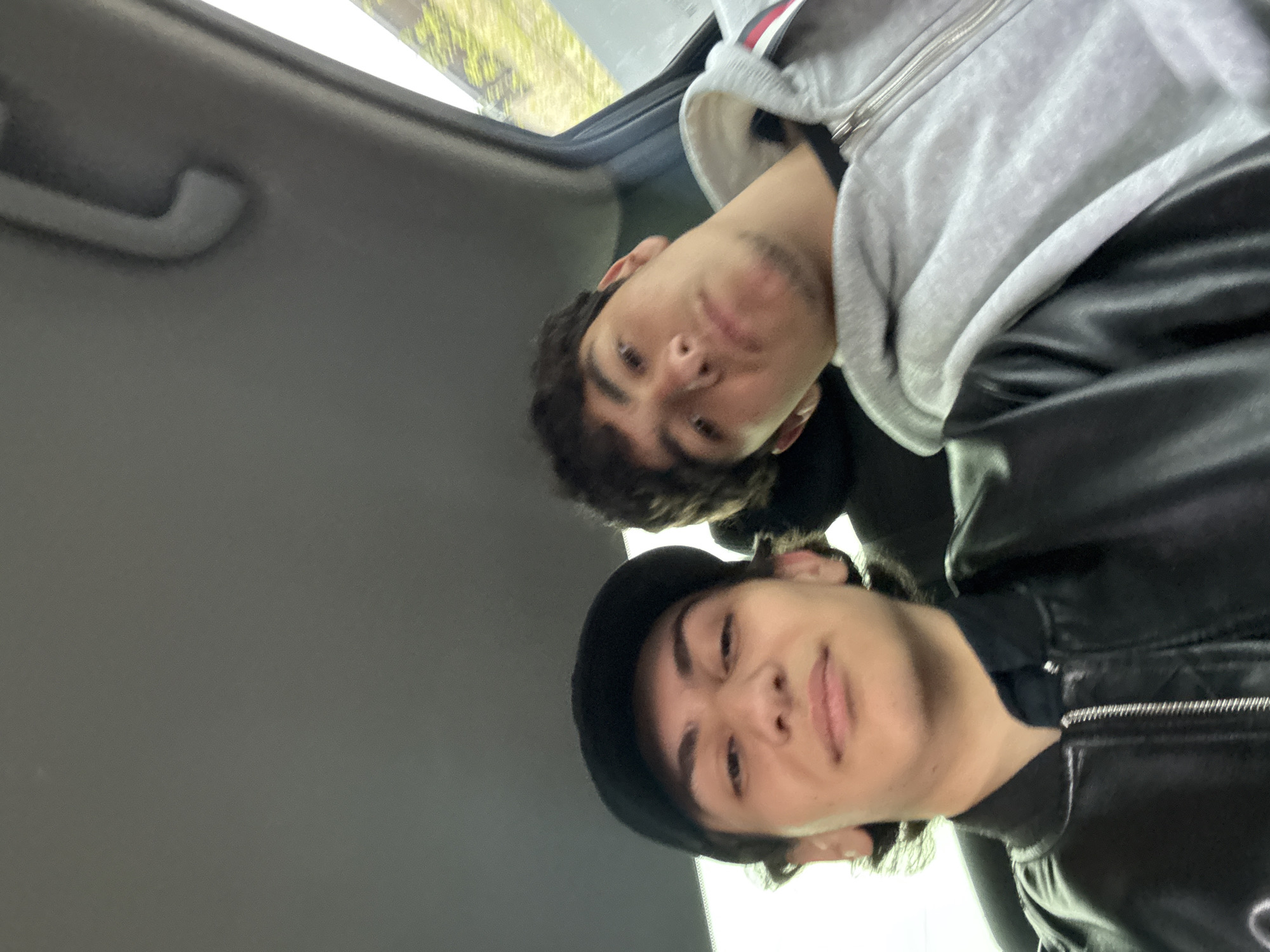Two young men are visible in the interior of a vehicle. The man in the foreground is wearing a black leather jacket and a dark-colored hat. The other individual, positioned slightly behind and above, is wearing a gray hooded sweatshirt. The setting is inside a car, indicated by the interior roof and handle visible in the frame. The image provides no location information about Yerevan, Armenia.