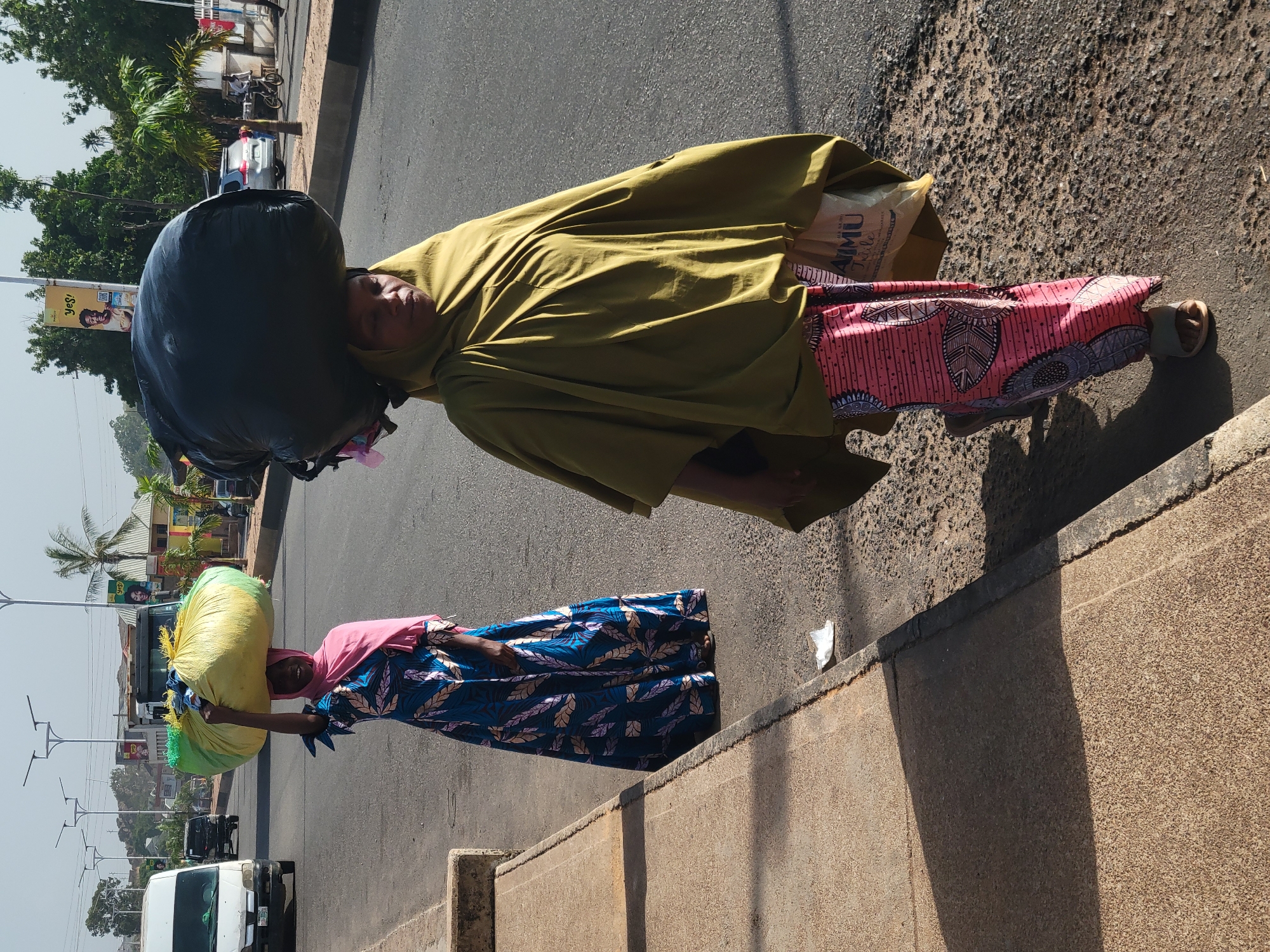 Two women carry loads on their heads along a street in Jos, Nigeria. One woman is on the sidewalk and carries a yellow bag while the other is on the road carrying a large black bag. The women wear long colorful dresses. The street is lined with buildings, poles, and trees. Cars are driving on the street.