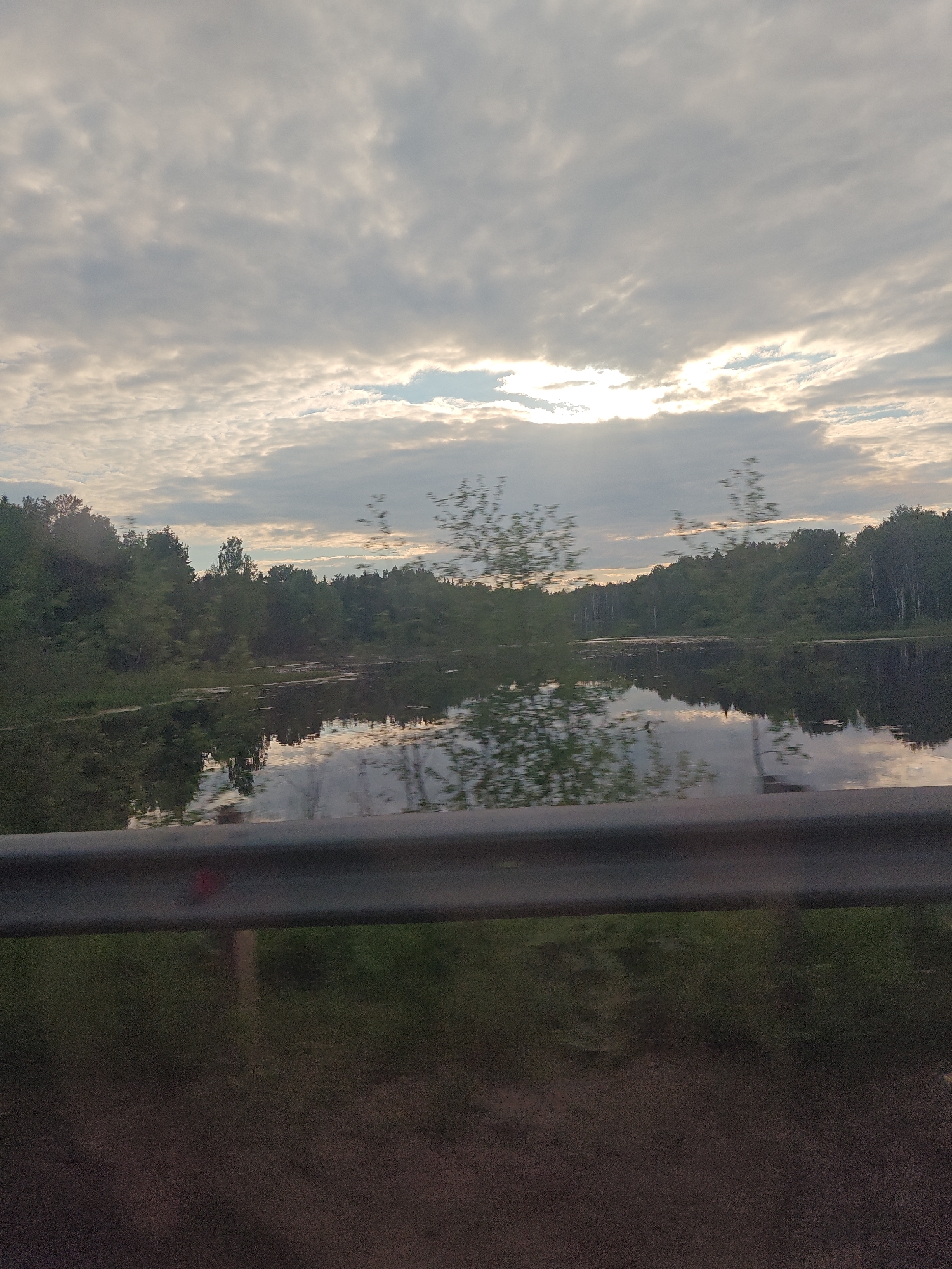 This image depicts a natural landscape, presumed to be near Kamennyi Kliuch, Russia. The scene is captured from a low vantage point, likely from a moving vehicle, with a dark grey metal guardrail occupying the lowest portion of the frame. Above the guardrail, a calm body of water, possibly a river or lake, reflects an overcast sky and the dense tree line on its far bank. The sky is largely covered by stratocumulus clouds, but a bright horizontal band of sunlight breaks through a gap slightly left of center, illuminating portions of the clouds with a soft, warm glow. The distant bank is covered by a lush, green deciduous forest, with reflections of the trees and sky visible on the water's surface. Small, dark patches of aquatic vegetation or debris are scattered across the water. The immediate foreground below the guardrail is dark and indistinct, with hints of green vegetation. No people, animals, or significant man-made structures beyond the guardrail are visible within the frame.