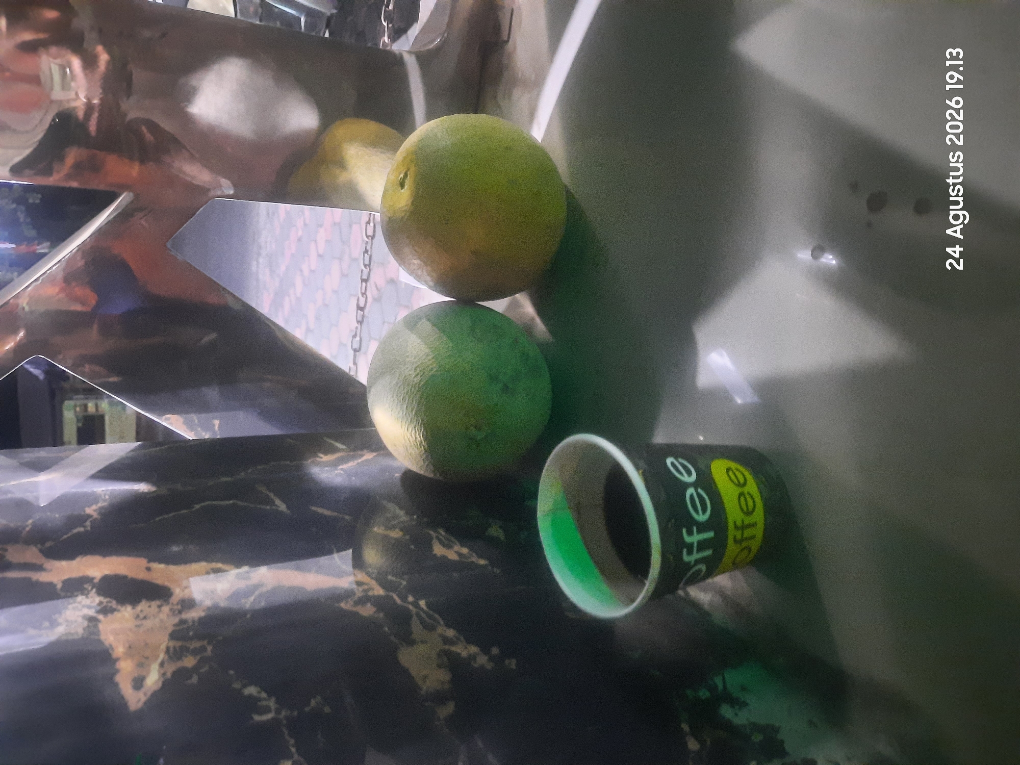 The image shows a close-up of a dark, marble-patterned cylindrical object on the left, and a reflective metallic sculpture on the right. In the foreground, a small paper cup with "coffee" written on it is filled with dark liquid. Two round fruits, one greenish and one yellowish, are placed on a flat surface between the cup and the metallic sculpture. The background appears to be an outdoor setting with patterned pavement and a chain, possibly indicating a public space in the city of Sukokerto, Indonesia. The timestamp "24 Agustus 2026 19.13" is visible in the lower right corner.