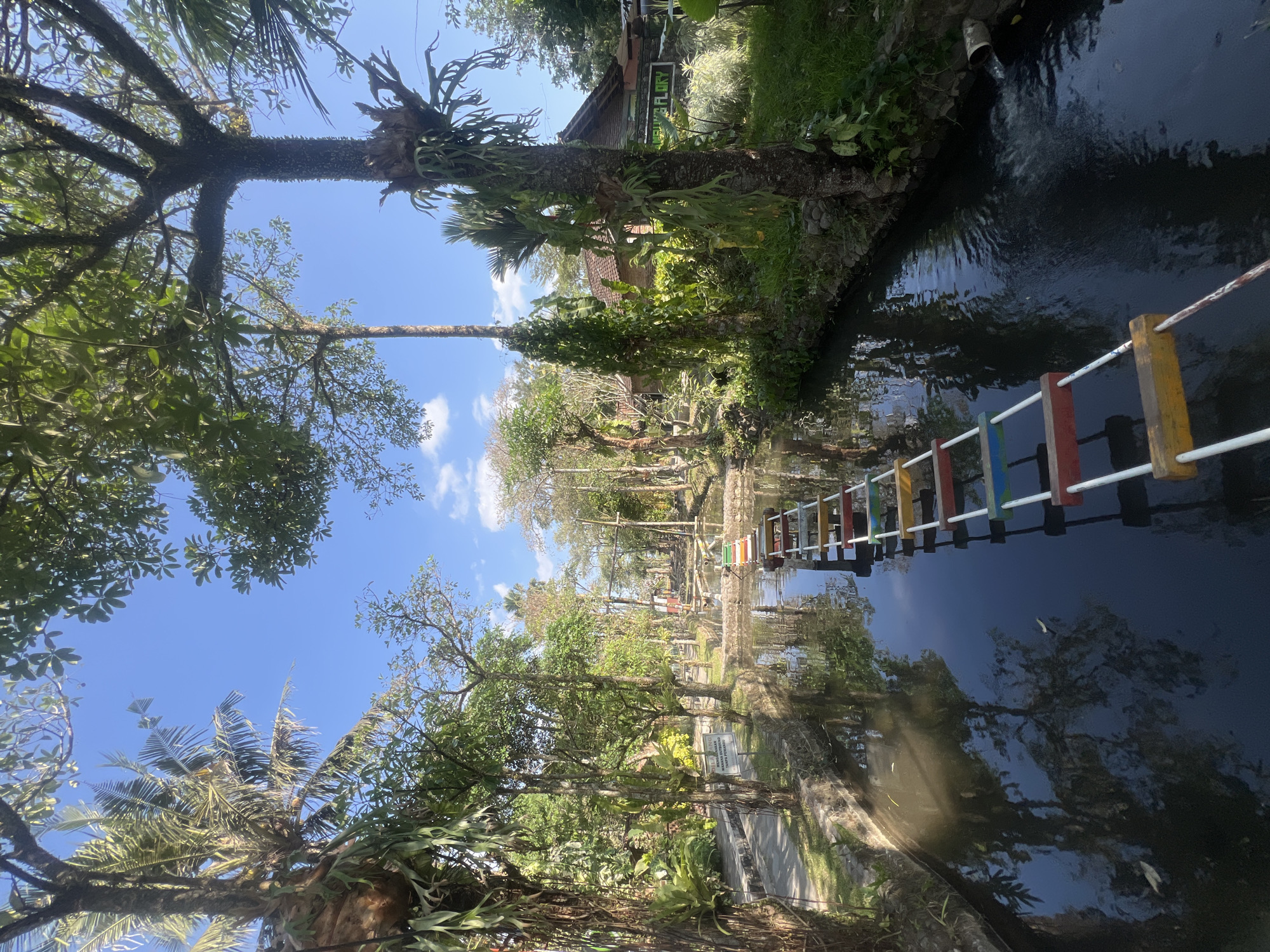 The image shows a picturesque canal with a rope bridge spanning across it. The canal is flanked by lush green vegetation and trees, with reflections of the trees and the blue sky with scattered clouds shimmering on the water's surface. The rope bridge has colorful wooden planks as steps, alternating in red, blue, and yellow. In the background, there are buildings with traditional architecture and more trees, suggesting a tranquil, possibly resort-like setting. A sign with text in an Indonesian language is visible on the left side of the canal. The scene appears to be a serene outdoor location in Tlogoadi, Indonesia, possibly a park or a botanical garden. There are no people visible in the image.