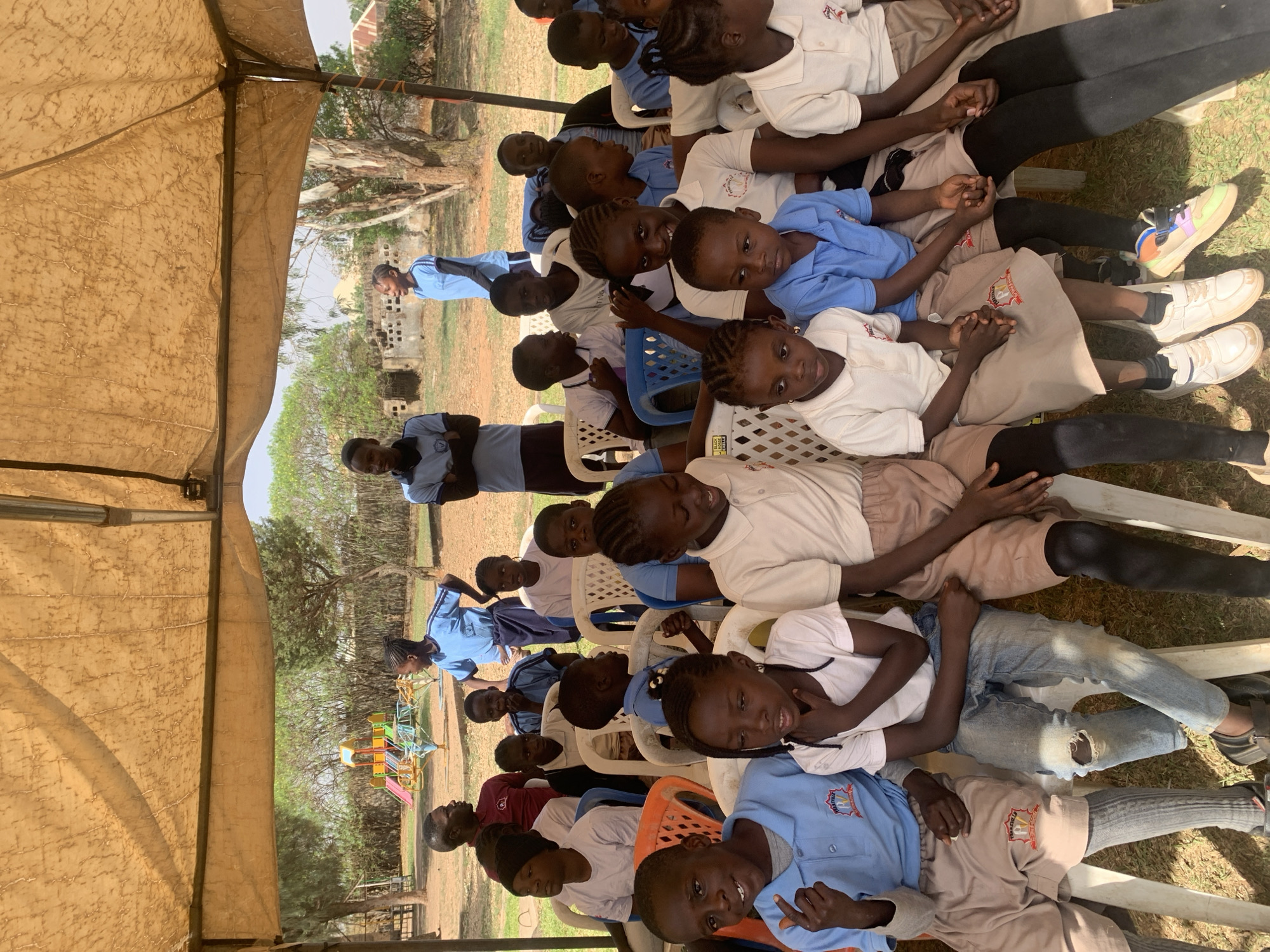 A medium shot captures a group of approximately twenty children and a few adults gathered outdoors under a large, tan-colored canopy or tent, providing shade from bright sunlight. The scene is set in what appears to be a schoolyard or community area in Jos, Nigeria, with playground equipment (swings and slides) and sparse trees visible in the background. Some simple concrete block structures are also seen in the distance.
The children, ranging in age from young primary schoolers to pre-teens, are seated on a mix of white, blue, and orange plastic chairs, arranged in rows. Many are wearing school uniforms: light blue polo shirts with dark trousers or khaki/beige skirts/shorts, and white polo shirts with similar bottoms. Some children wear more casual attire, including jeans and darker shirts. One child in a white shirt has a black patch on their arm with the text "BLACK BELT" partially visible. School crests or logos are visible on the uniforms of several children; one clearly shows "GREAT BEGINNERS SCHOOLS".
The children are mostly looking towards the camera, with some smiling, while others have curious or neutral expressions. A few adults, also in uniform-like blue shirts, are visible in the background, standing. The overall mood appears calm and organized, suggesting a gathering, perhaps a class or an event. Bright daylight indicates it's likely midday or early afternoon. The ground is dry and dusty.