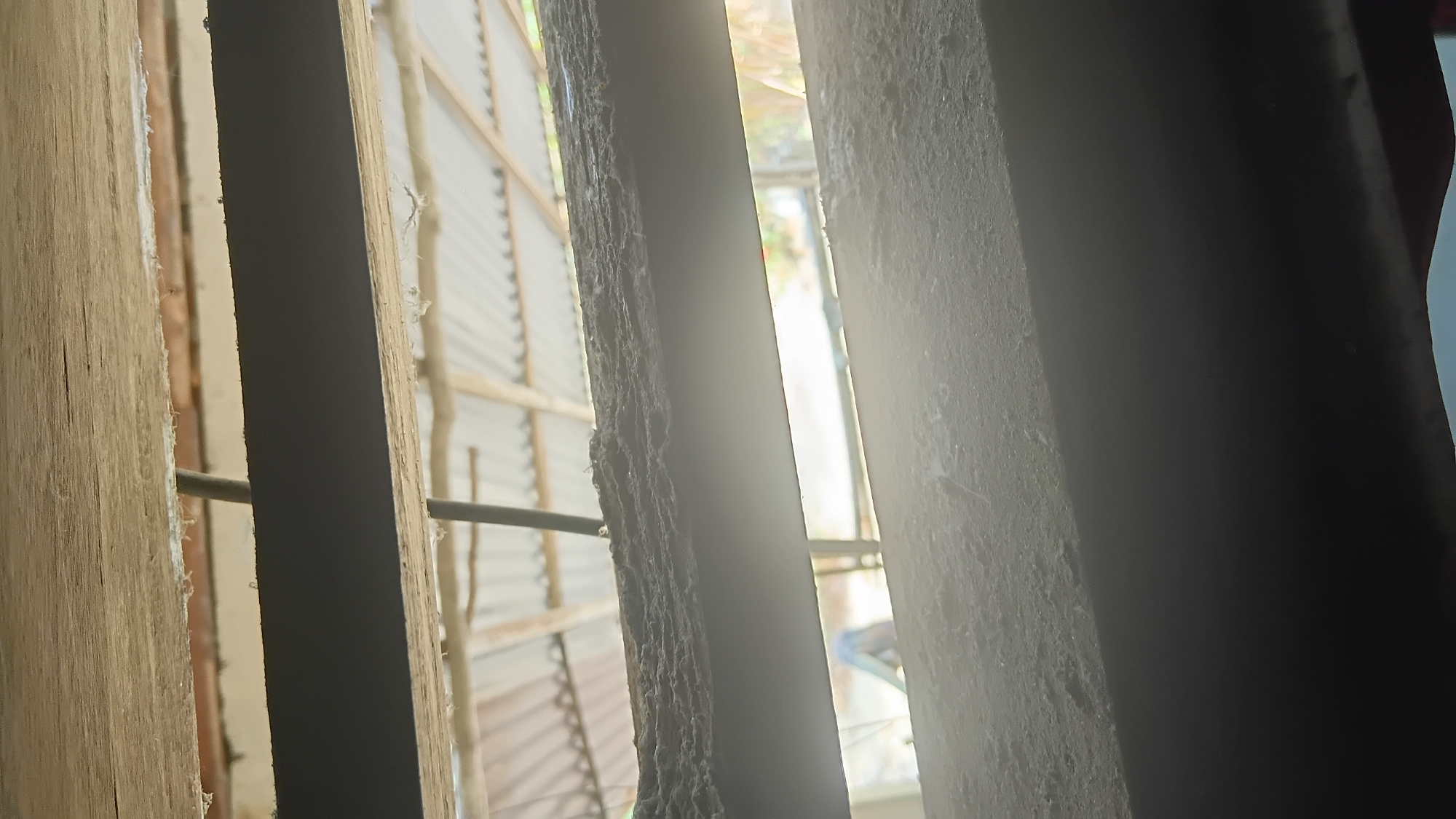 The image depicts a view through what appears to be louvers or slats, suggesting a window or vent. The material of the slats seems to be a combination of wood and possibly metal or painted material, with a significant accumulation of dust and grime on their surfaces. Through the gaps, a corrugated metal roof structure is visible, indicating an outdoor or semi-outdoor setting. Sunlight filters through, creating bright areas and shadows. The environment appears to be rustic and possibly exposed to the elements. The image does not provide enough information to determine the specific location within Prabumulih, Indonesia, beyond suggesting a location with such construction. No people or specific interactions are discernible.