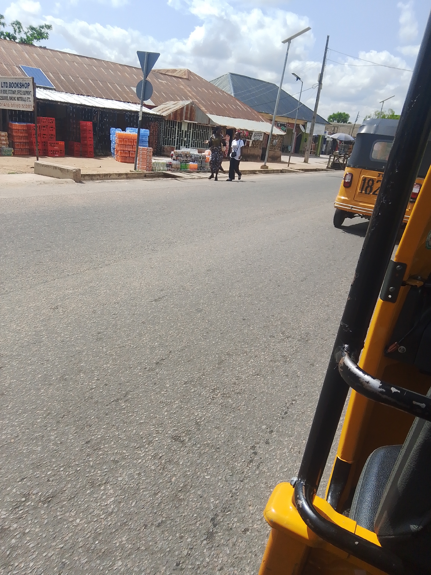 This media file captures a street scene in Jalingo, Nigeria, likely during daytime with a mix of sun and clouds. The foreground is dominated by the yellow and black metal frame of a tricycle, suggesting the photo is taken from the perspective of its passenger or driver.
In the background, a row of businesses lines the street. One shop, identified by text as "LTD. BOOKSHOP," displays stacks of orange and blue plastic crates, likely holding beverages or other goods. The shop's roof is corrugated metal. Other shops are visible, with one appearing to be a "DEPOT." A yellow tricycle with the number "182" visible on its back is parked further down the street.
Several people are walking on the sidewalk in front of the shops. Two individuals are walking together, one in a patterned dress and the other in a dark shirt and pants. Another person in a white shirt and dark pants is walking in the opposite direction. The overall atmosphere appears to be that of a typical, bustling market or commercial area. Streetlights and utility poles are also visible. The presence of the tricycles and the architecture of the buildings suggest a tropical or sub-tropical climate.