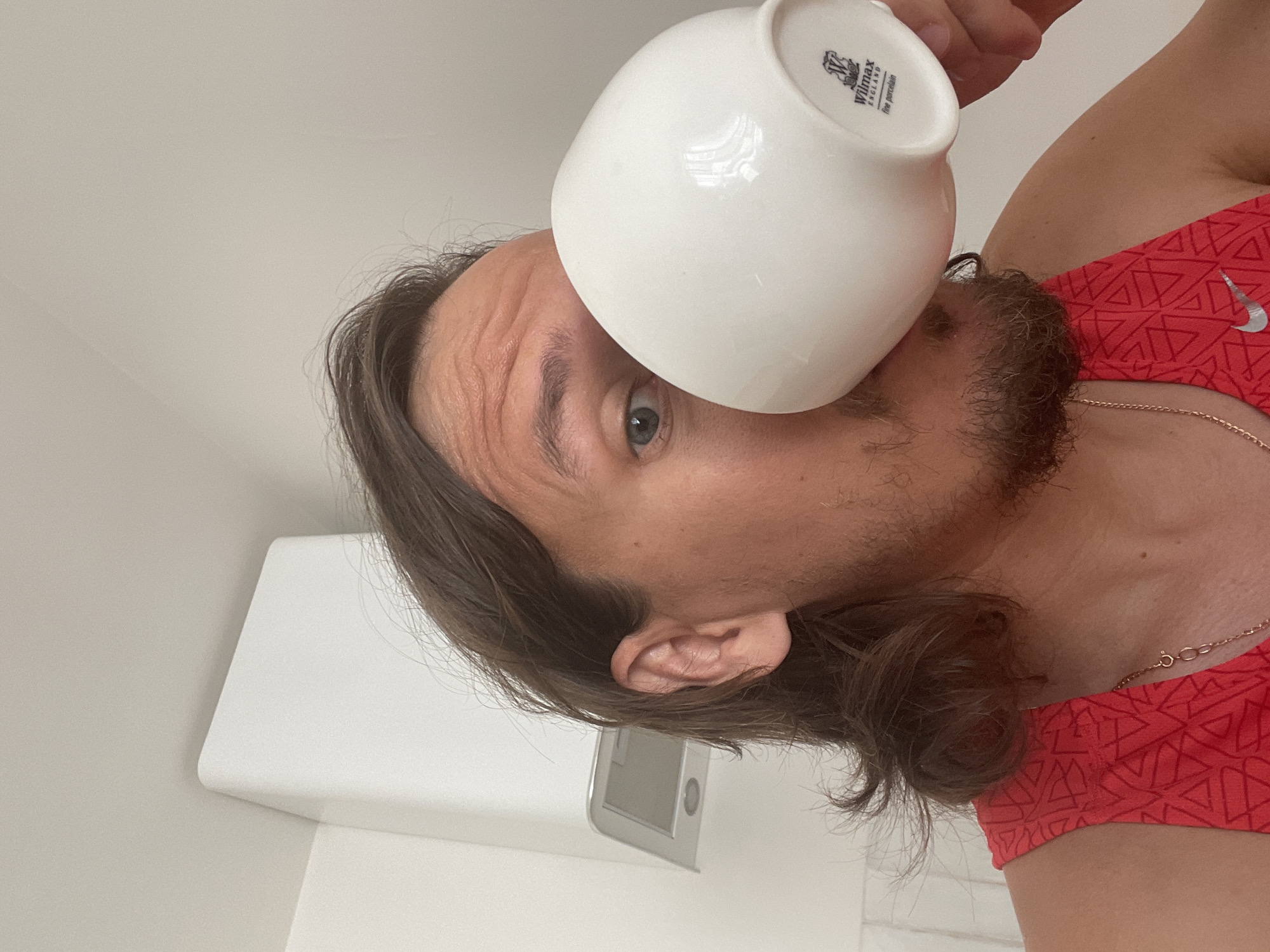 A man with long brown hair and a beard is drinking from a white Wilmax England mug. He is wearing a red tank top with a Nike logo. The background is a white wall with a white object, possibly a shelf, attached to it. The image provides no evidence to suggest the location is Izhevsk, Russia.