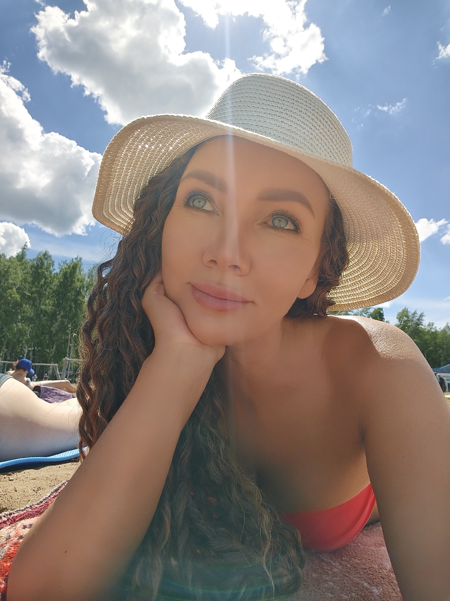 A woman is lying on a beach, wearing a wide-brimmed straw hat and a red bikini top. She is resting her head on her hand and looking up at the sky. The sun is shining brightly, with visible lens flare effects. The sky is blue with fluffy white clouds. Behind her, trees and some beachgoers can be seen in the distance, suggesting a recreational area. The woman has long, curly brown hair and striking green eyes. She appears relaxed and content. The overall impression is of a sunny day at the beach.