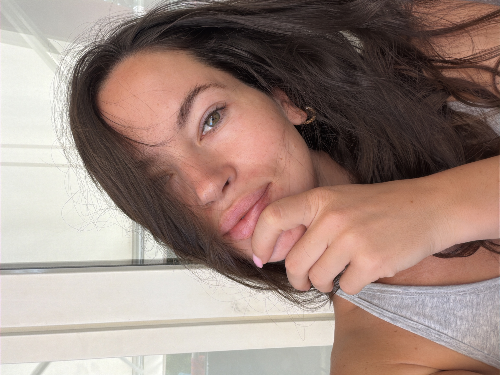 A woman with long, dark, wavy hair is pictured in a close-up shot. Her hair is partially obscuring her face. She is wearing a light gray tank top. Her right hand is raised to her face, with her fingers touching her lips. Her left eye is visible and appears to be green. She is wearing a small gold hoop earring in her left ear. The background appears to be indoors, with vertical white panels that could be part of a building or door frame. The lighting suggests it is daytime. There are no discernible landmarks or features that specifically place the image in Salou, Spain.