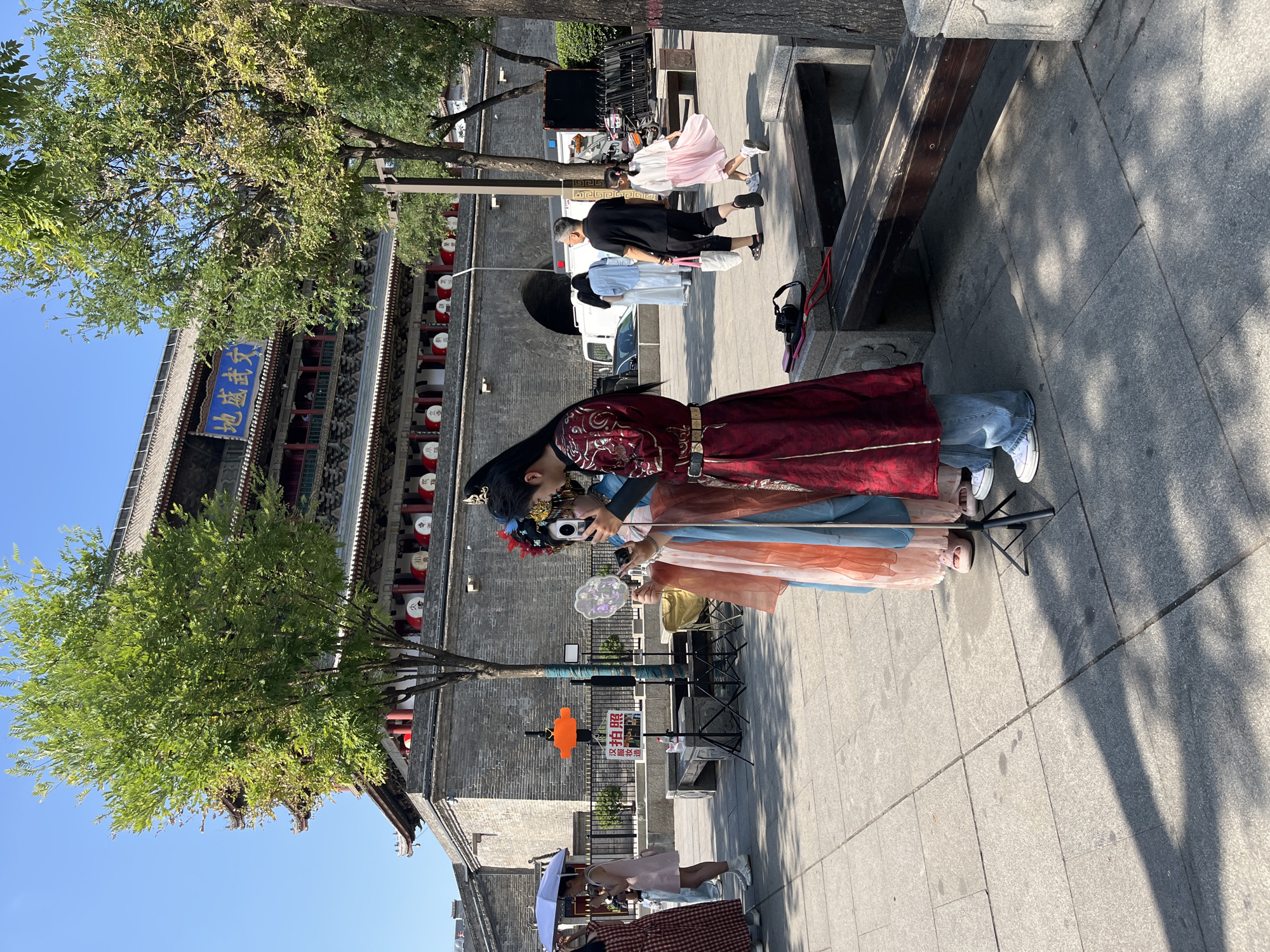 In an outdoor plaza, in front of a large, ancient-style building with a tiered roof and a prominent blue sign bearing Chinese characters, two individuals dressed in traditional Chinese attire are taking photos of each other with smartphones. The person on the right wears a vibrant red and gold robe with intricate embroidery, and has their hair styled with ornate accessories. The person on the left wears a lighter colored robe with flowing sleeves. A fan is held by the person on the left. A camera is visible on a tripod in the foreground, suggesting a photo-taking session. Several other people are visible in the background, some walking and others standing, with a white van and a motorbike also present. Trees with green foliage are scattered throughout the scene, casting shadows on the paved ground. The sky is clear and blue, indicating a sunny day. The architectural style of the building suggests it is a historical landmark, possibly a gate or a temple.