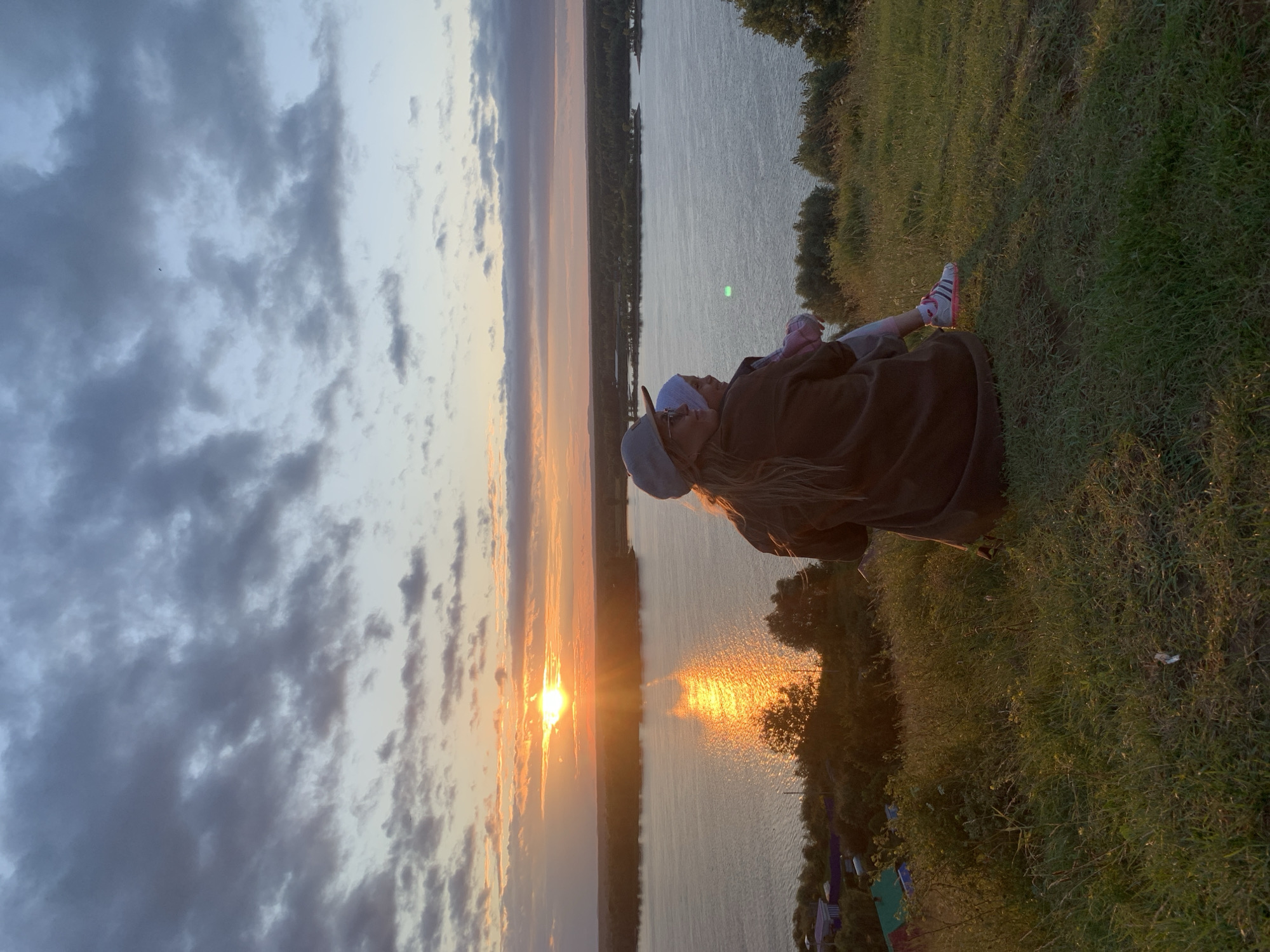 The image depicts a woman and a child from behind, sitting on a grassy hill overlooking a wide river at sunset. The sky is filled with dramatic clouds, tinged with the orange and yellow hues of the setting sun, which casts a shimmering reflection on the water. In the distance, a treeline and a small settlement are visible on the opposite bank of the river. The woman is wearing a cap and a brown jacket, and the child, whom she is holding, is wearing a white hat. The overall scene suggests a moment of quiet observation and connection amidst a beautiful natural landscape. The location is identified as Olkhovka, Russia.