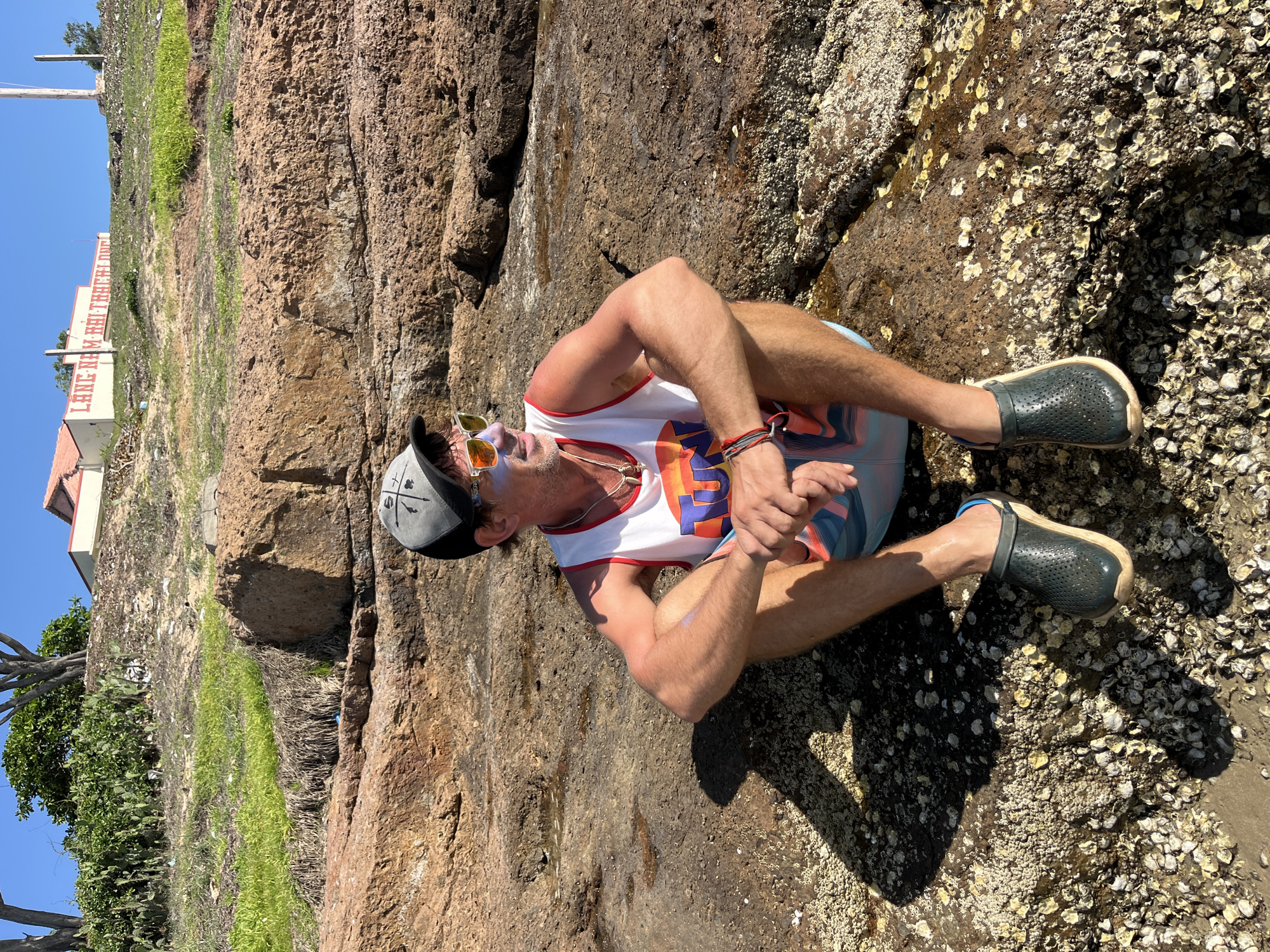 A man with sunglasses and a baseball cap is sitting on a rocky, barnacle-covered shoreline, facing the ocean. He is wearing a white tank top with colorful graphics and patterned shorts. His hands are clasped in front of him. The sun is bright, casting shadows on the rocks, suggesting it is daytime.
In the background, a steep, rocky cliff rises, with patches of green grass and scrub vegetation. To the far left, a building with "LANG HAM HUI KHOI KHANH" written in red letters on its side is visible, along with some trees. The overall scene suggests a warm, sunny day at a coastal location. The man appears relaxed, enjoying the scenery. The weathered rocks and the presence of barnacles indicate a tidal area.