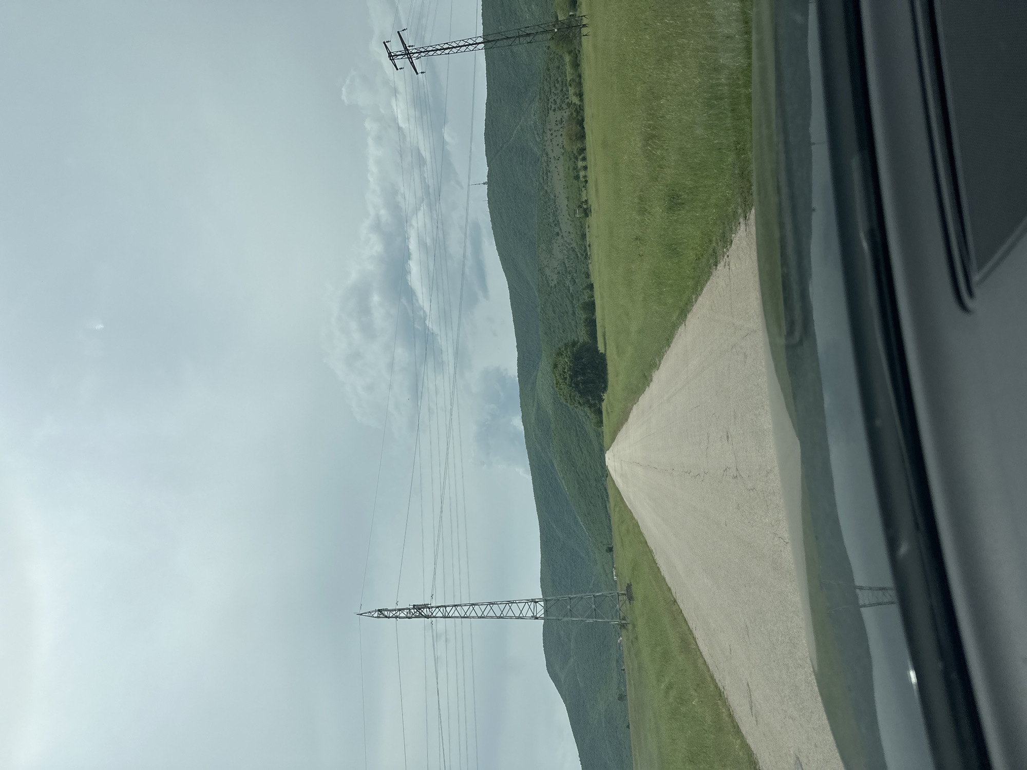 The image shows a view from inside a vehicle in Gara Oreshets, Bulgaria. The foreground is dominated by the interior of the vehicle on the right side of the frame. Outside, a straight, paved road stretches into the distance, flanked by a green grassy field on one side and a tree-covered hillside on the other. Two electrical transmission towers with power lines are visible against a cloudy sky.
