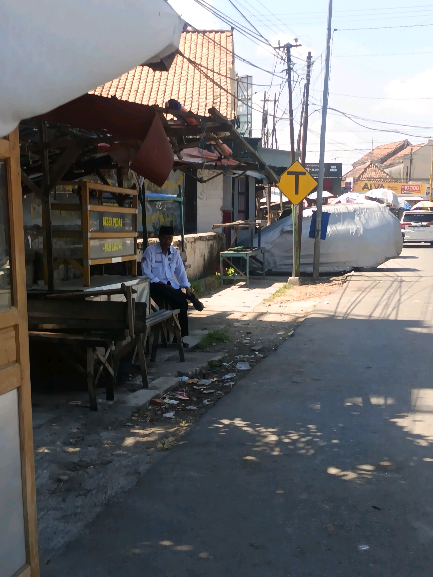 This image depicts a street scene in Blega, Indonesia, characterized by its informal commercial stalls and a residential backdrop. In the foreground, a man wearing a white long-sleeved shirt and black pants, with a black cap, is seated on a wooden bench beside a stall. The stall has a wooden frame and glass panels, with signage indicating "BEBEK PEDAS" (Spicy Duck) and "NASI JAGUNG" (Corn Rice). The ground around the stall is unpaved and littered with debris.
To the right of the stall, a large object covered in a light-colored tarp is prominently visible. A yellow traffic sign with a black "T" symbol is mounted on a pole in front of it. Beyond the tarp, a white car is parked on the street.
In the background, traditional Indonesian rooftops with terracotta tiles are visible, interspersed with utility poles and a network of electrical wires. The sky is clear and blue. The overall impression is one of a developing urban environment with elements of both commerce and everyday life.