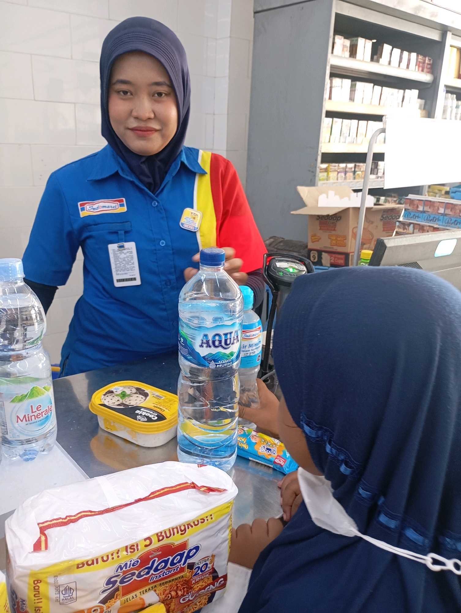 In a retail store in Babelan, Indonesia, a cashier wearing a blue Indomaret uniform with red and yellow accents and a dark blue hijab stands behind a counter. She is looking at the camera with a slight smile. In front of her on the counter are two bottles of water, one labeled "Le Minerale" and the other "Aqua," an ice cream container labeled "Classic Creamy Cookie Creamy," and a bag of "Mie Sedap Instant" noodles. To the right of the cashier, partially obscured by a dark blue hijab, is another person, presumably a customer, reaching for something on the counter. A barcode scanner is visible on the counter. In the background, shelves are stocked with various products. The environment appears to be typical of a convenience store or minimarket.