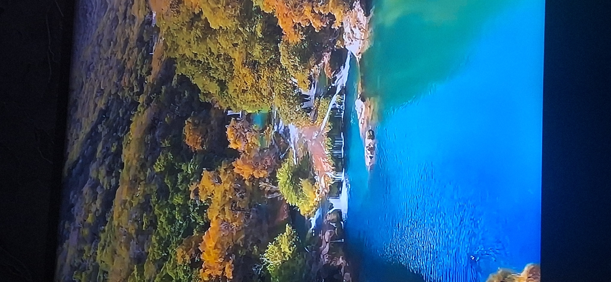 The image depicts a vibrant, multi-tiered waterfall cascading into a series of bright blue pools. The surrounding landscape is lush and autumnal, with trees displaying a spectrum of green, yellow, and orange foliage. Rocky outcrops punctuate the water's edge, and a thin layer of mist rises from the falls, catching the light. The water in the pools is exceptionally clear, revealing submerged rocks and creating a striking contrast with the verdant vegetation. There are no visible people or man-made structures in the immediate vicinity of the waterfall. The scene is natural and serene, characterized by the dynamic flow of water and the rich colors of the fall season. The image does not contain any identifiable elements that would place it in the city of Bila Tserkva, Ukraine.