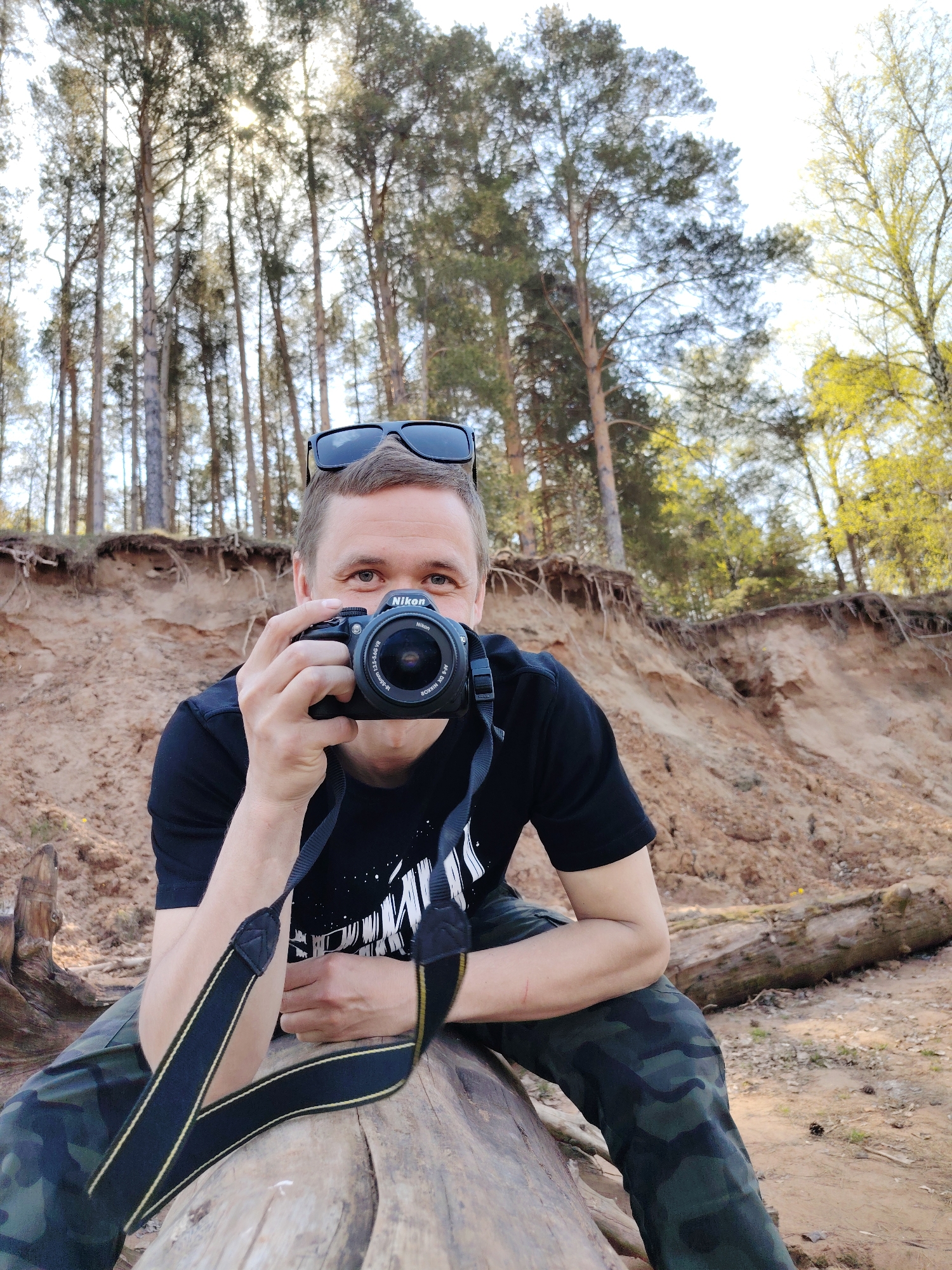 A man is crouched on a fallen tree trunk in what appears to be a forest area in Chaikovsky, Russia. He is holding a Nikon camera to his face, as if taking a picture. The man is wearing a black t-shirt, camouflage pants, and has sunglasses perched on his head. A camera strap is visible around his neck. Behind him is a sandy embankment, with tall trees in the background that appear to be pines and birches. The lighting suggests it is daytime.