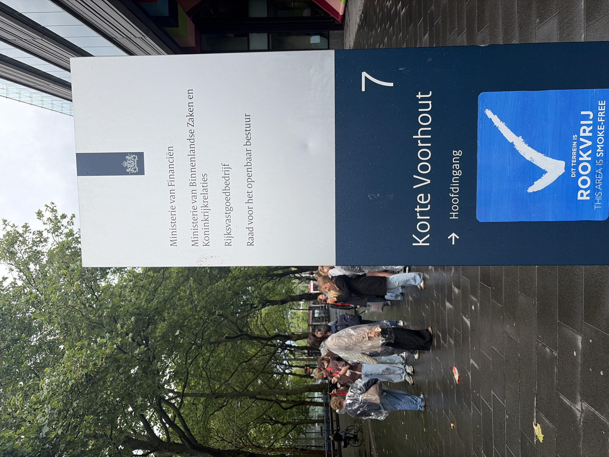 This outdoor image, taken in The Hague, Netherlands, features a prominent informational sign on an overcast day, with clear indications of recent rainfall. The ground is wet, and several individuals in the mid-ground are wearing transparent rain ponchos.
The sign, composed of a white upper section and a dark blue lower section, lists various Dutch government bodies in black text: "Ministerie van Financiën" (Ministry of Finance), "Ministerie van Binnenlandse Zaken en Koninkrijkrelaties" (Ministry of Interior and Kingdom Relations), "Rijksvastgoedbedrijf" (Central Government Real Estate Agency), and "Raad voor het openbaar bestuur" (Council for Public Administration).
The dark blue portion of the sign displays "Korte Voorhout," a street name, followed by the number "7." Below this, an arrow points to "Hoofdingang" (Main Entrance). Additionally, a blue square sticker is affixed to the sign, featuring a white checkmark logo and the text "DIT TERREIN IS ROOKVRIJ" (THIS AREA IS SMOKE-FREE), with the English translation provided underneath.
A group of people, some equipped with rain gear, are gathered on the wet, paved surface beneath a line of green trees. Their activity appears to be standing or waiting. A modern building with a reflective glass facade is partially visible on the left side of the frame.