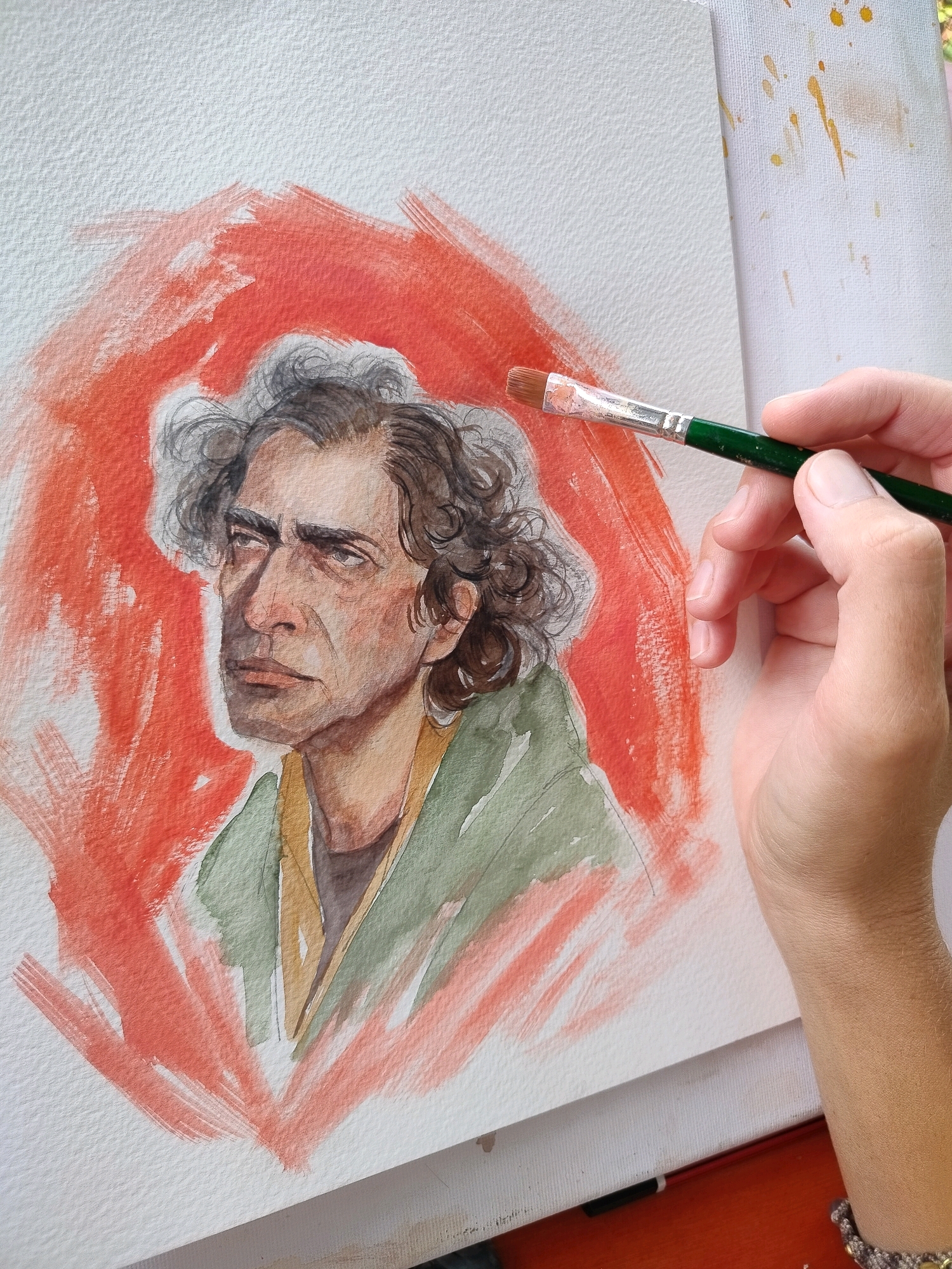 The image shows a close-up of a watercolor painting in progress, depicting a man's face with curly dark hair and a pensive expression, set against a background of bold red brushstrokes. A hand holding a paintbrush is positioned on the right side of the frame, actively working on the painting. The painting is on textured white paper, which is part of a larger art piece or canvas. The overall scene suggests an artist at work in what appears to be an indoor setting, possibly a studio. There is no indication of the city of Gagra, Abkhazia, or any other specific geographical location.
