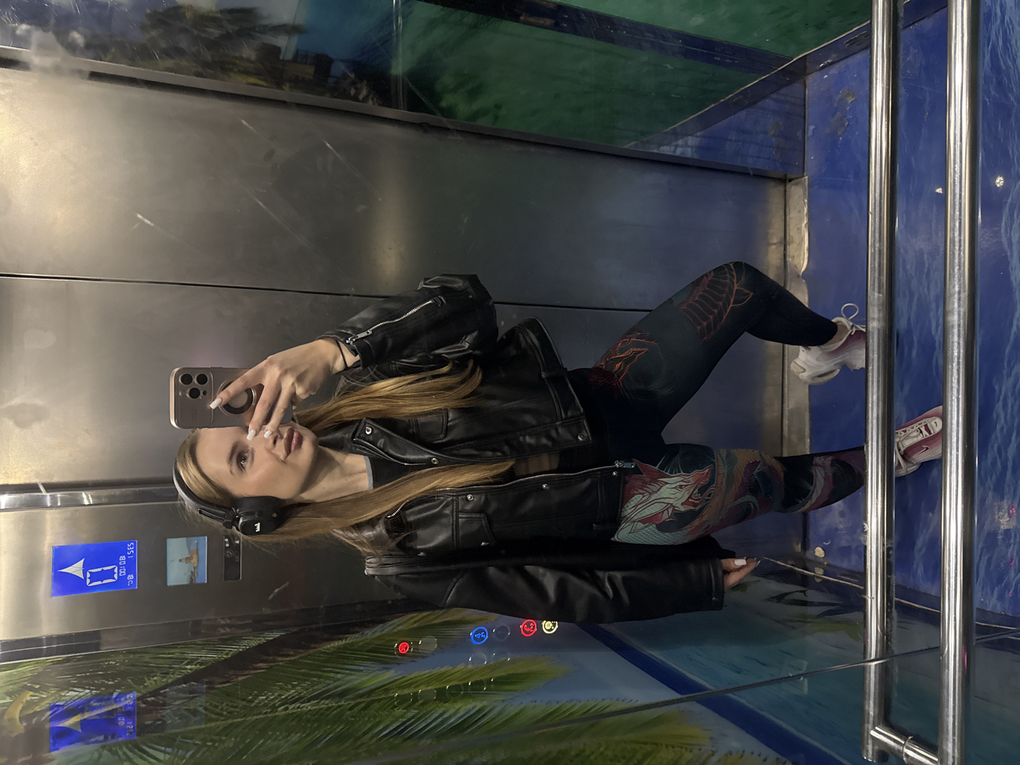 A young woman is taking a selfie in an elevator. The elevator has a mural of a tropical scene on the walls. The woman is wearing a black leather jacket, patterned leggings, and sneakers. She is also wearing headphones. The location is Budva, Montenegro.