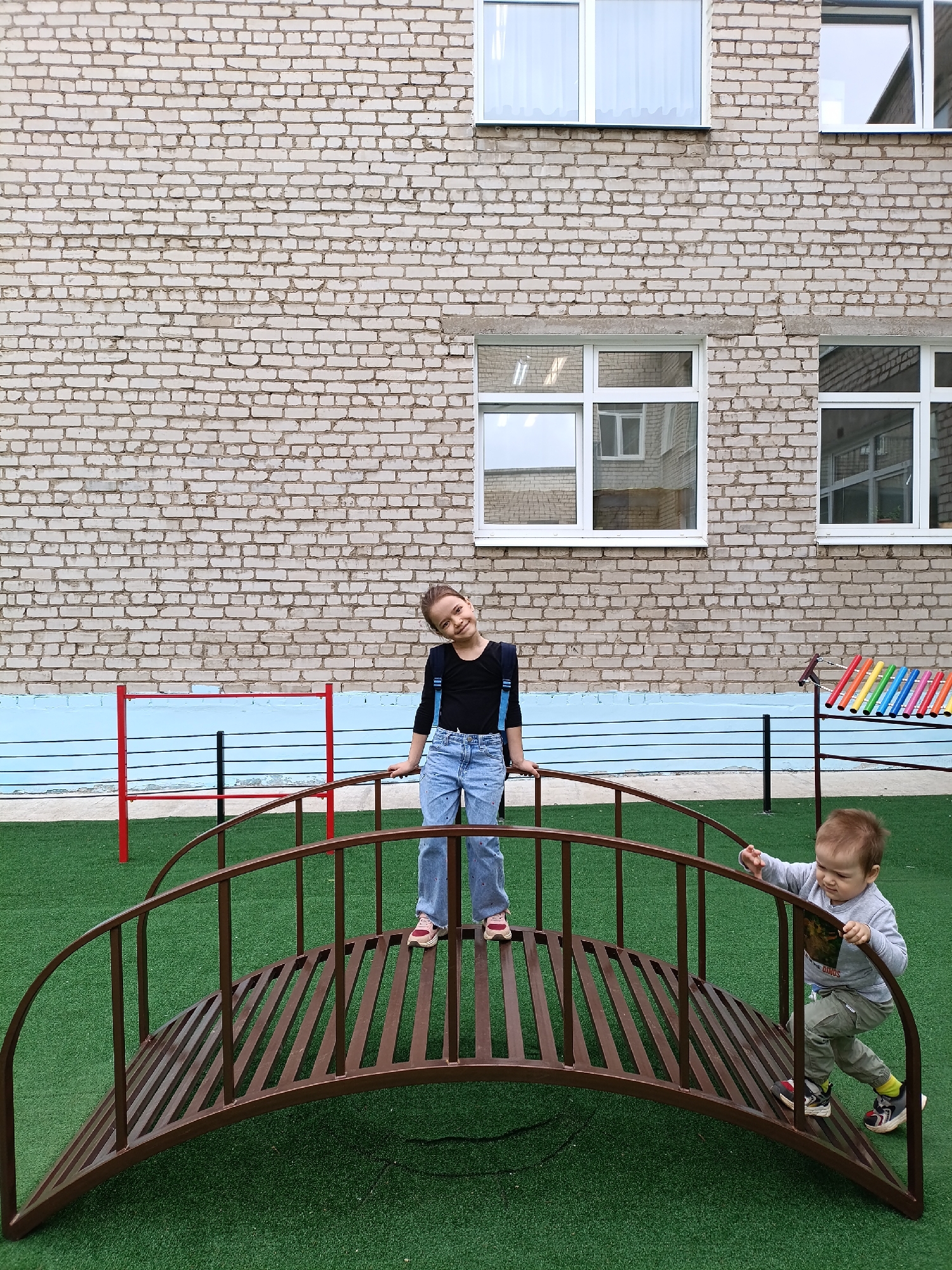The image depicts a playground scene in Chaikovsky, Russia. A girl with dark hair, wearing a black long-sleeved shirt, light blue jeans with dark blue suspenders, and pink shoes, stands on the top of a brown metal bridge-shaped structure. To the right of the girl, a toddler wearing a grey long-sleeved shirt and olive green pants with colorful sneakers is descending the bridge. The playground has green artificial turf. Behind the bridge is a light tan brick building with three windows. To the left of the girl is a red, black, and white goalpost, and to the right is a colorful xylophone. A light blue painted surface borders the base of the brick building. The sky is overcast.