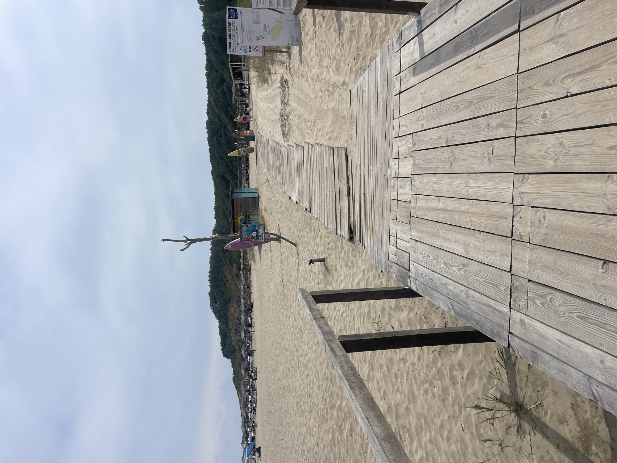The image depicts a wooden boardwalk leading across a sandy beach in Rezovo, Bulgaria. The boardwalk is made of weathered wooden planks and has a rustic railing on the left side. The beach is covered in light brown sand, with a few tufts of dry grass growing near the boardwalk. In the distance, numerous umbrellas and lounge chairs are set up on the beach, indicating a populated area. Behind the beach, a line of green trees forms a backdrop against a slightly overcast sky. A signpost with information in Cyrillic script is visible to the right of the boardwalk, near the beach's edge. Surfboards are also present, leaning against a small wooden structure. A few people are seen scattered across the beach, enjoying the seaside.
