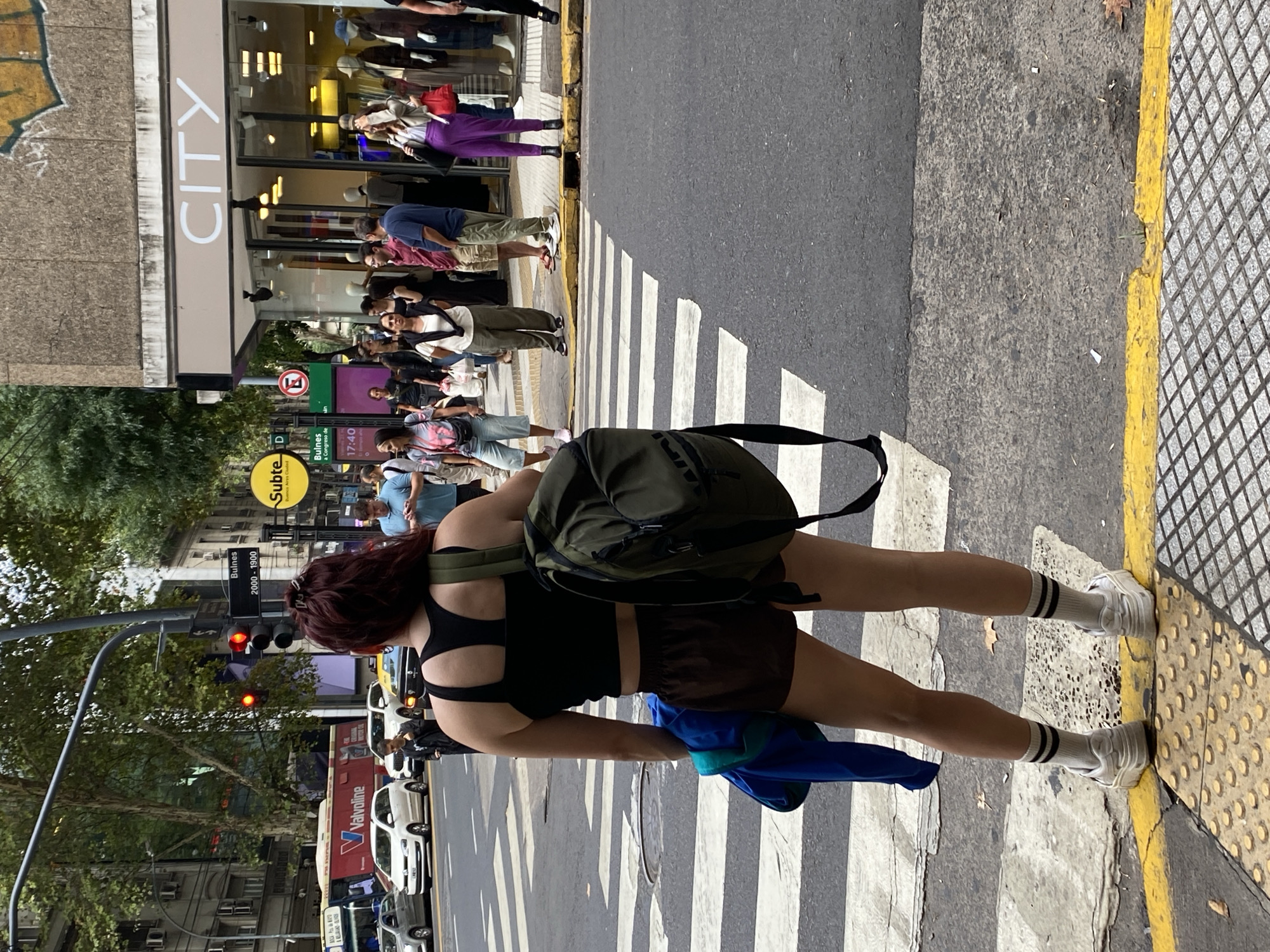 The image captures a street scene in Buenos Aires, Argentina, at the intersection of Bulnes street, indicated by a street sign displaying "Bulnes 2000 - 1900" and a yellow circular "Subte" sign. A digital clock sign shows "Viernes 23 de Enero 17:40."
In the foreground, a person is viewed from behind, standing on the sidewalk at the edge of a pedestrian crosswalk. This individual has reddish-brown hair, wears a black athletic tank top, brown shorts, white socks with two black stripes, and white platform sneakers. An olive-green backpack is carried over their left shoulder, and a folded blue jacket is held in their left hand. The sidewalk beneath them features gray tactile paving with raised circular patterns, bordering a yellow-painted curb line.
The asphalt street ahead contains a marked pedestrian crosswalk. Multiple vehicles are visible, including several white cars, a yellow taxi, and a red public bus with "Valvoline" branding further down the street. Traffic lights for cross traffic are red.
On the opposite sidewalk, approximately ten to fifteen pedestrians are present, some walking and others standing near the entrance of a store labeled "CITY." One person is wearing purple trousers and a light-colored jacket, holding a red handbag. Another individual in a light blue shirt is looking at a mobile phone. Buildings with multiple floors line both sides of the street, and trees with green foliage are visible. Small pieces of debris and dried leaves are scattered on the ground surfaces.