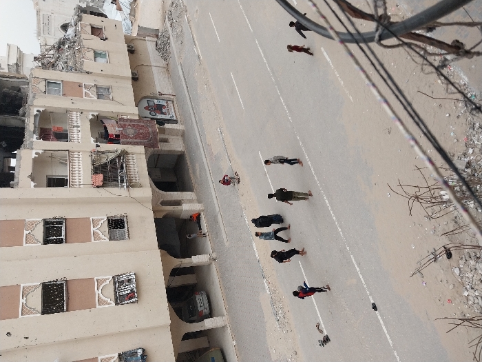 The image presents an aerial view, severely tilted, looking down at a damaged urban street scene during what appears to be midday due to the short shadows. The perspective is from a high vantage point, possibly a higher floor of a building or a drone, with power lines or cables partially obscuring the upper right corner of the frame.
On the left side of the image, a multi-story building, likely residential, shows significant damage. The upper floors are particularly affected, with large sections of walls missing, exposing interior spaces and twisted rebar. Windows are either broken, boarded up, or entirely absent, leaving gaping holes. A prominent red patterned rug hangs from a balcony railing on one of the middle floors, while another patterned textile, possibly a rug or blanket, hangs from a lower balcony. The lower floors of the building appear more intact, featuring arched entryways and windows. A dark-colored car, possibly a sedan or small SUV, is parked beneath one of these archways on the ground level. Above this, on the building's facade, there's a small billboard or sign depicting a figure, possibly a cartoon character, with a prominent red element on its head, but no readable text is clearly visible.
Running diagonally across the image, separating the damaged building from an unpaved area, is a paved road. White lines are painted on its surface, indicating lanes. Several individuals are scattered along this road, appearing to walk or stand. They are viewed from above, making their faces indistinct. From left to right, near the building, one person in a red shirt and dark pants is visible. Further along the road, a group of approximately six individuals is seen. They are dressed in casual clothing, mostly dark tops and pants, with one person wearing red pants. Their postures suggest casual movement or pauses.
To the right of the paved road, there is an unpaved, sandy or dirt-covered area that runs parallel to the road. This area is strewn with various types of debris, including what looks like twisted metal, broken concrete, plastic bags, and general refuse, suggesting a neglected or war-affected environment. The power lines in the foreground, consisting of multiple dark cables, descend from the top right, adding to the visual impression of an active but possibly neglected infrastructure.
Overall, the scene depicts everyday life continuing amidst visible structural damage and debris, suggesting a challenging urban environment, possibly in a conflict zone or an area affected by natural disaster or severe urban decay.